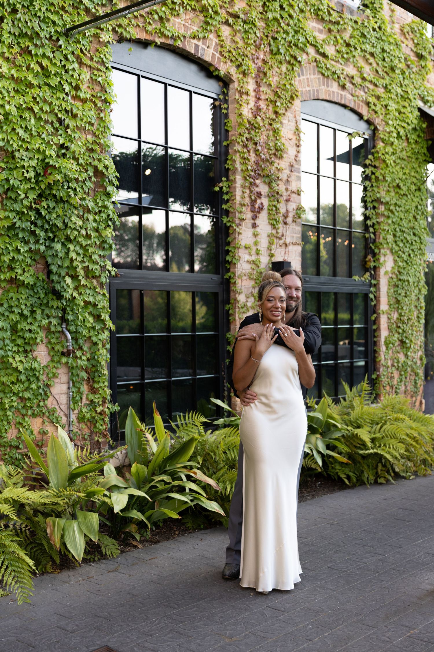 Couple posing outdoors by ivy-covered brick building, woman in white satin slip dress, man hugging her from behind.