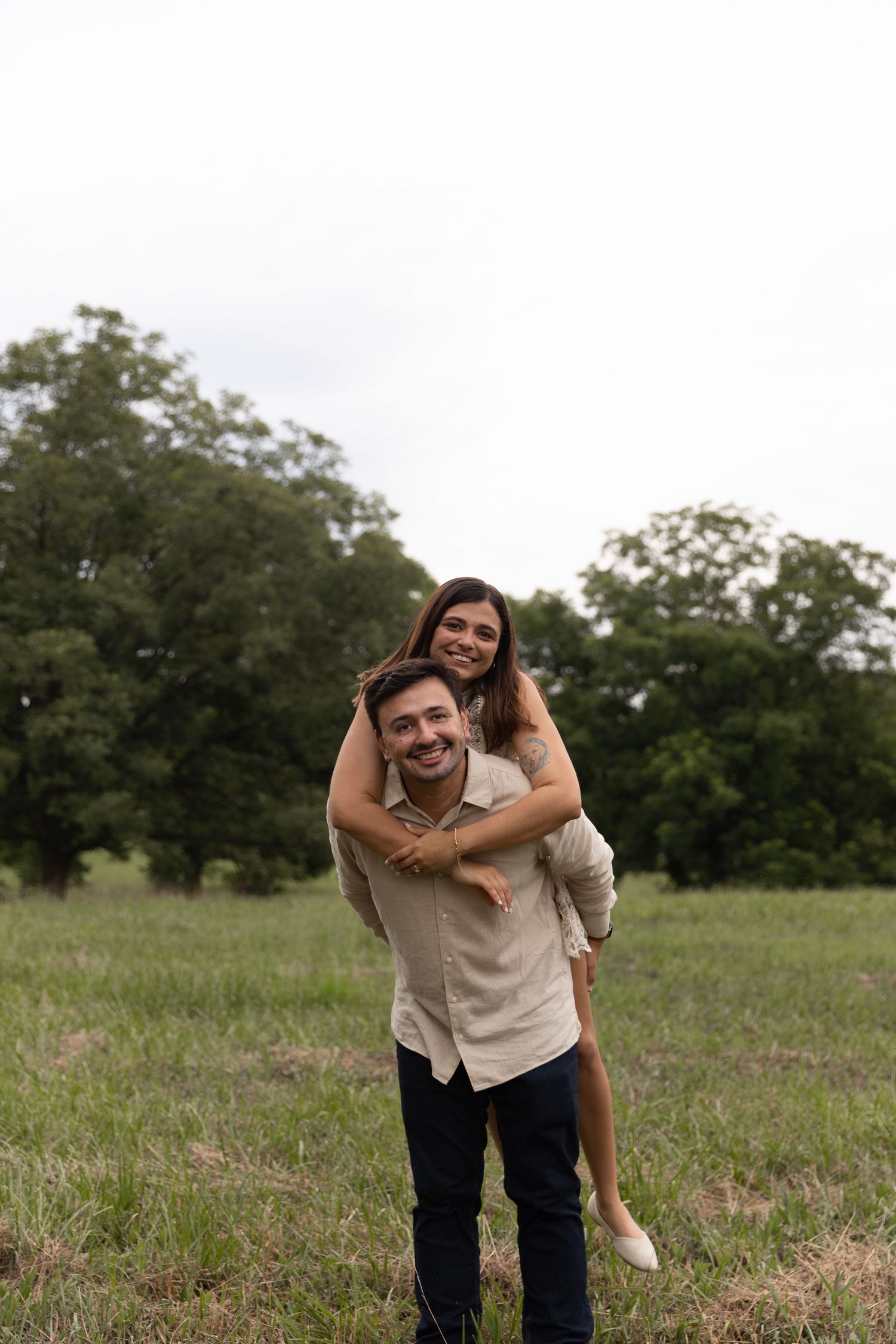 Happy couple in green field, woman riding piggyback on smiling man, trees in background.