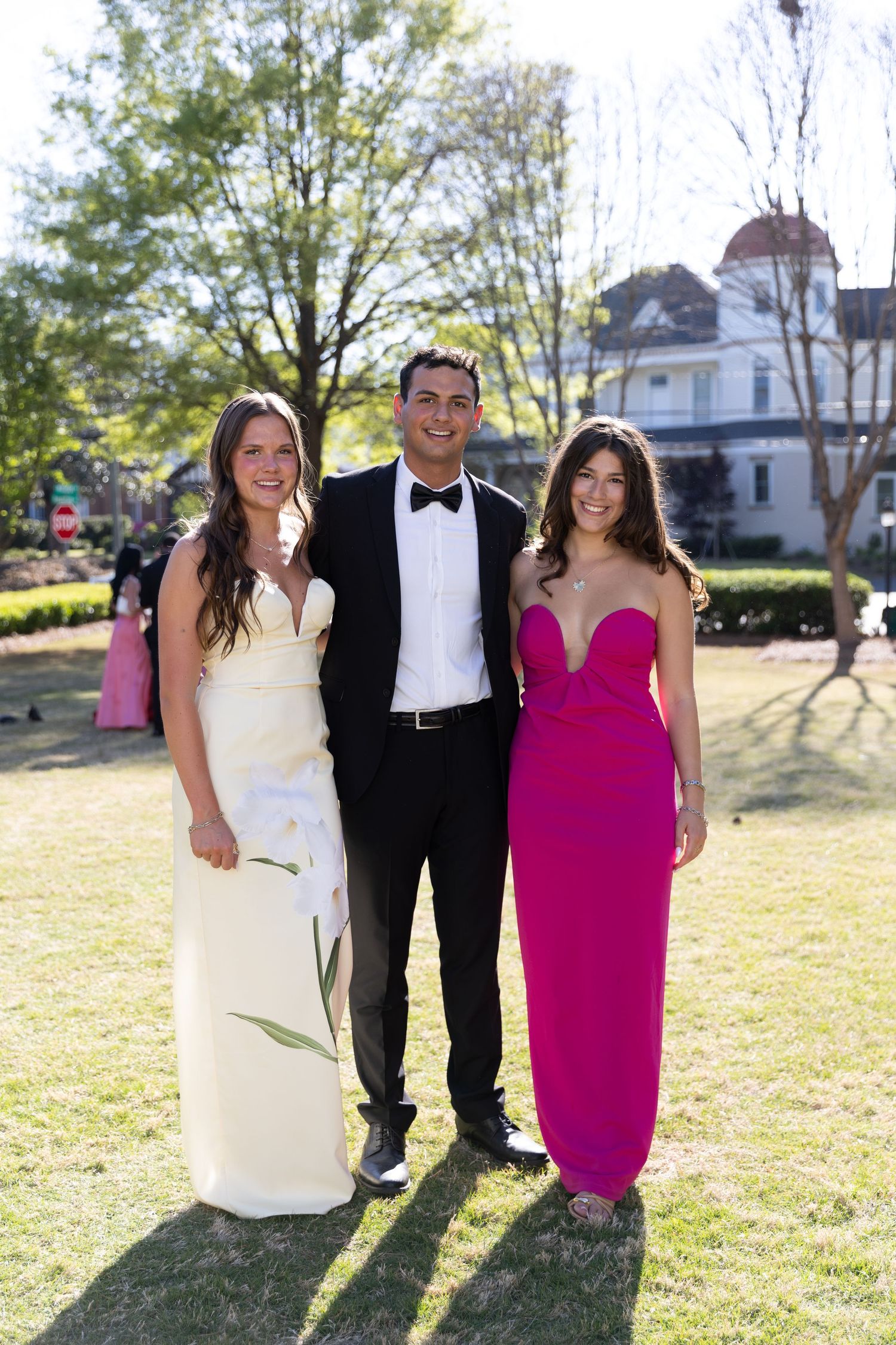 Three young adults in formal wear posing outdoors, two women in gowns flanking a man in tuxedo.