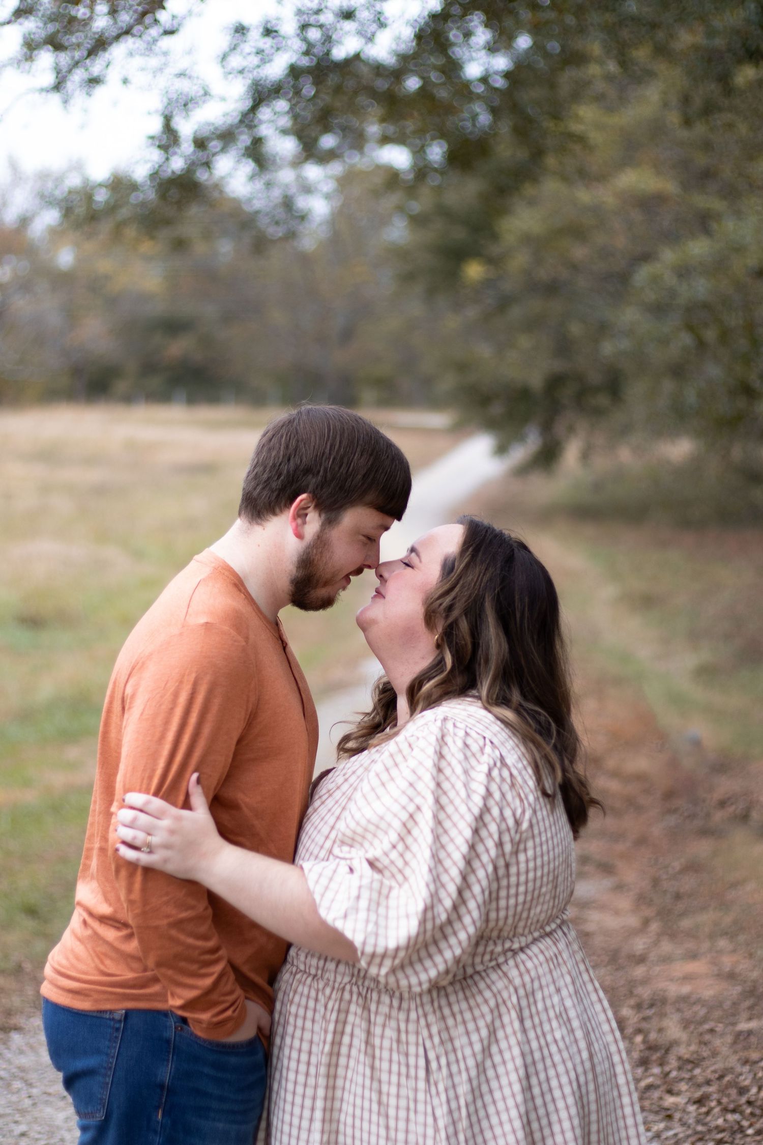 A couple shares a romantic nose-to-nose moment outdoors, the man in orange and woman in plaid dress embracing on a nature path.