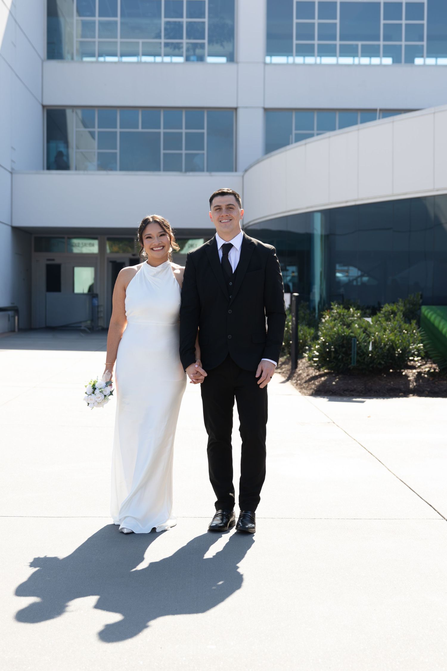 Smiling bride and groom holding hands outside a modern building on their wedding day.