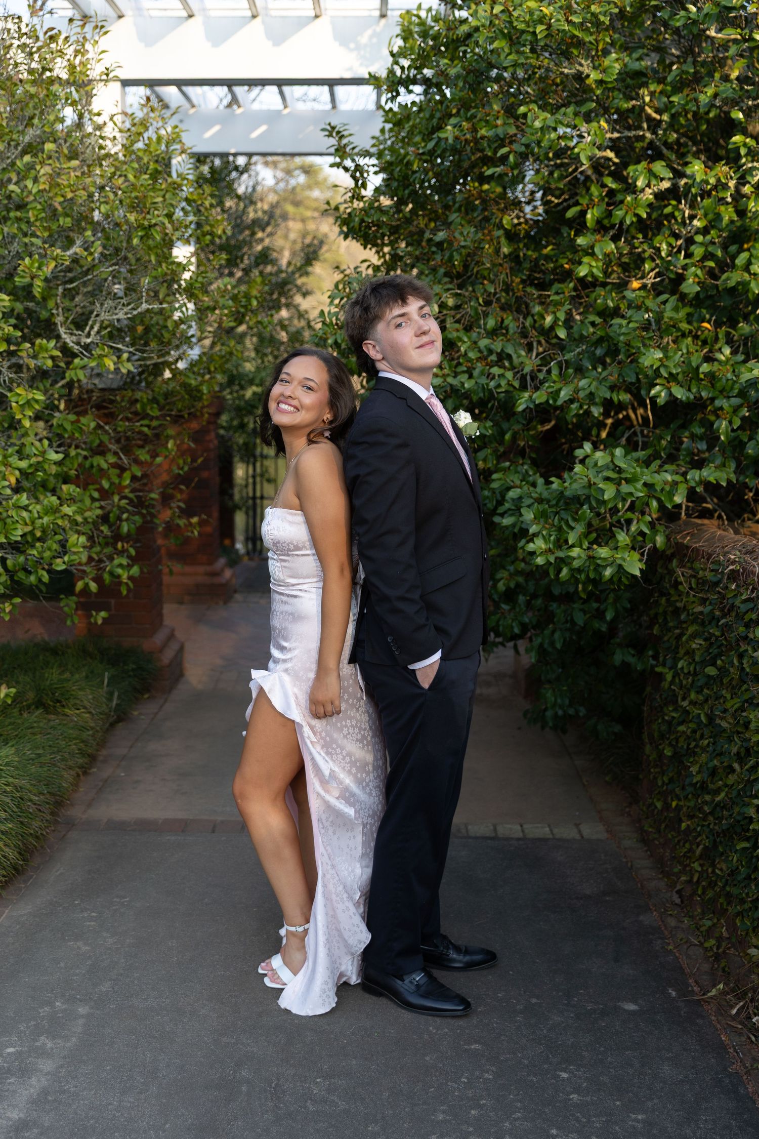 A young couple poses back-to-back in formal prom attire outdoors, she in a white gown, he in a black suit.