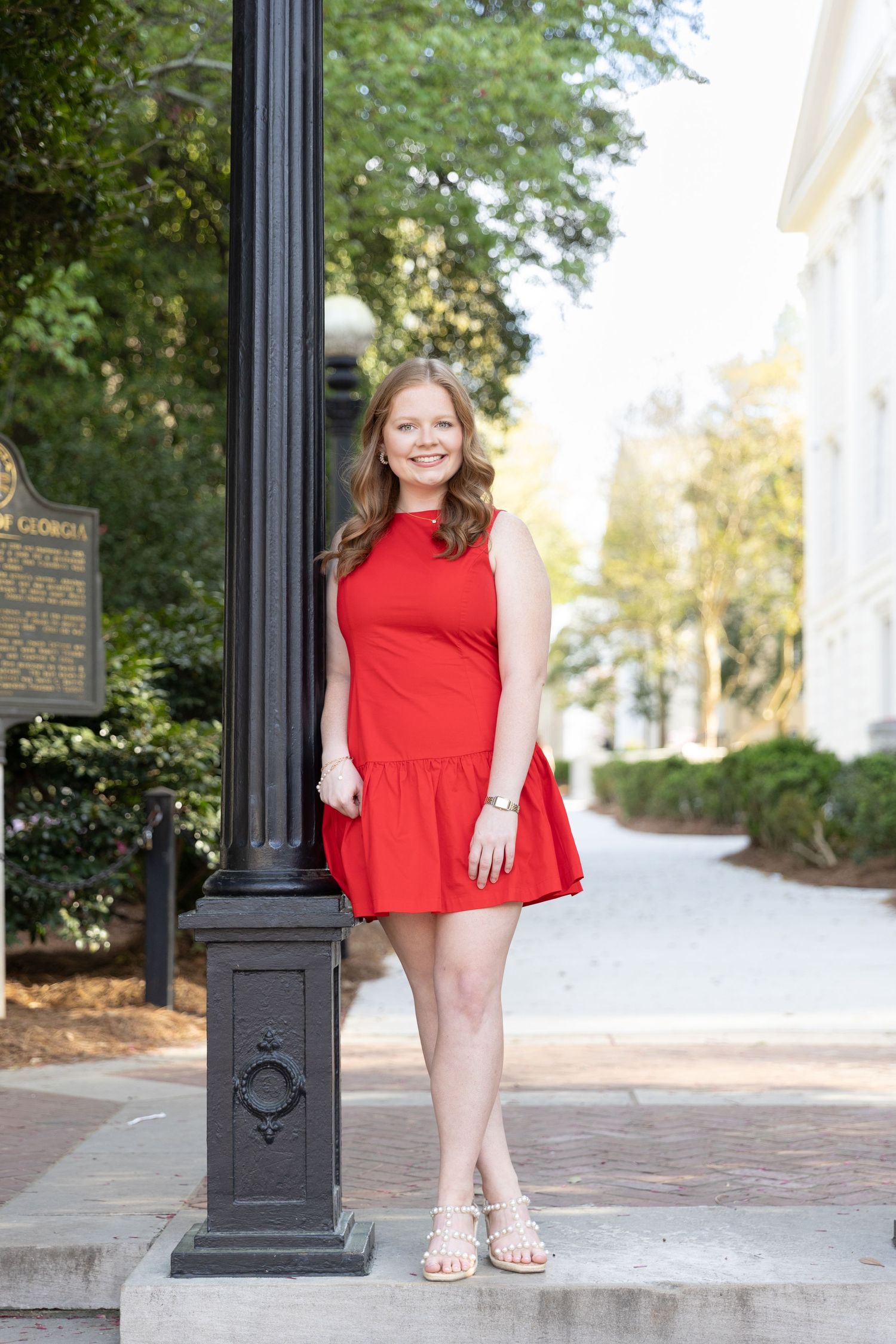 Young woman in red sleeveless mini dress posing by a black lamppost on a sunny outdoor walkway.