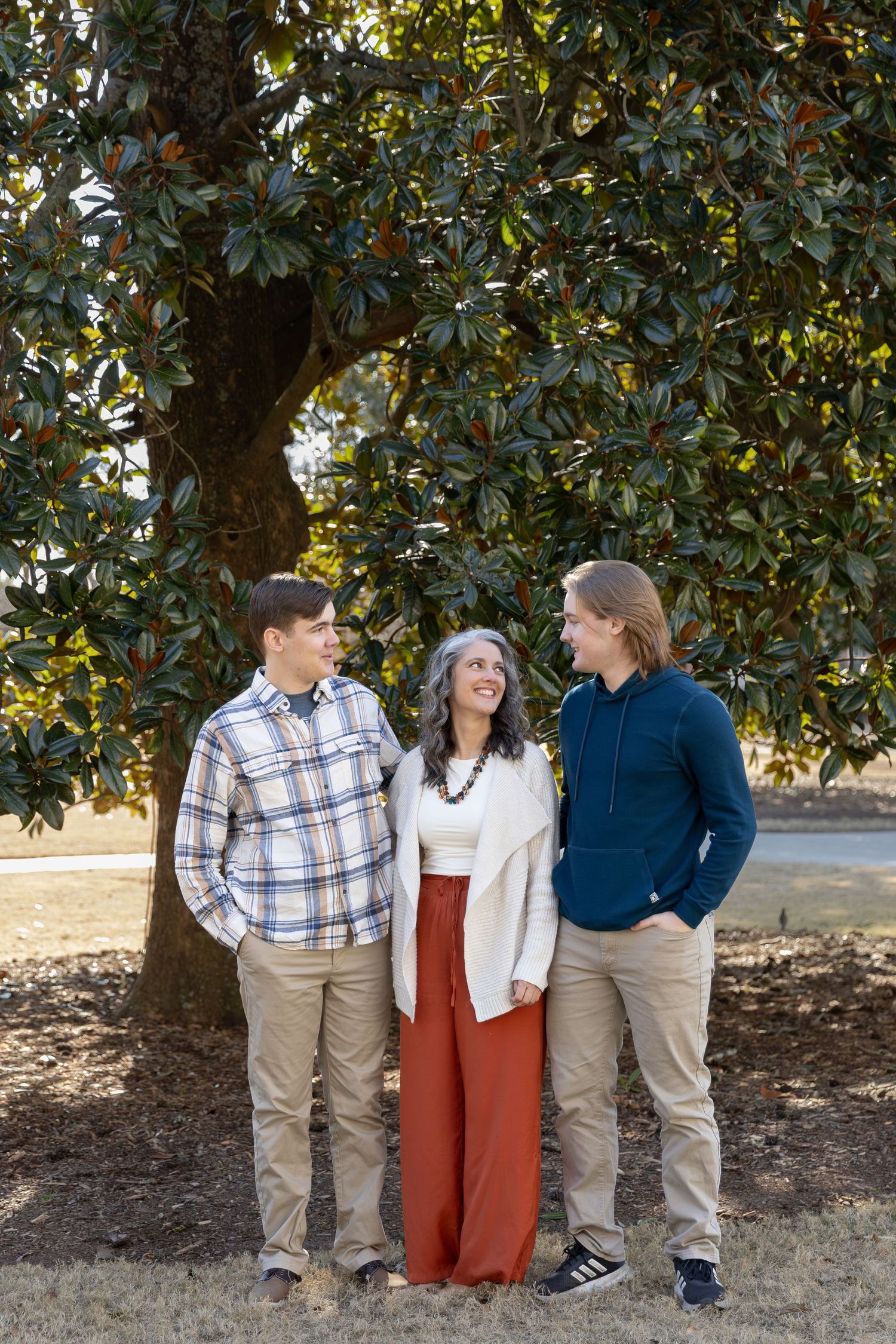 A woman with gray hair stands between two young men under a magnolia tree, all smiling warmly in casual attire.