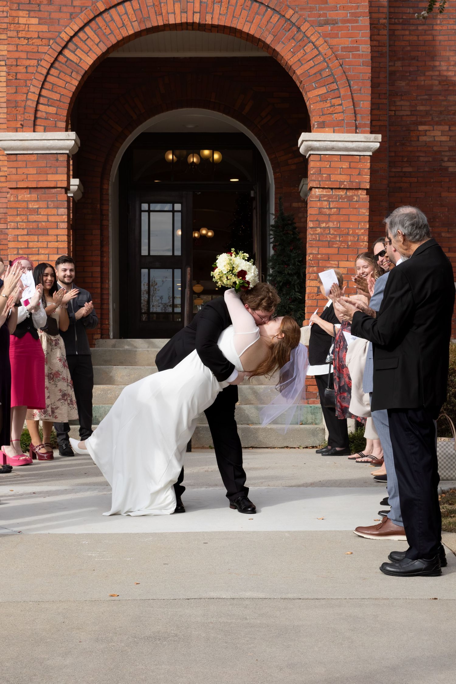Groom dips bride for a kiss outside a red brick church as wedding guests cheer and applaud.