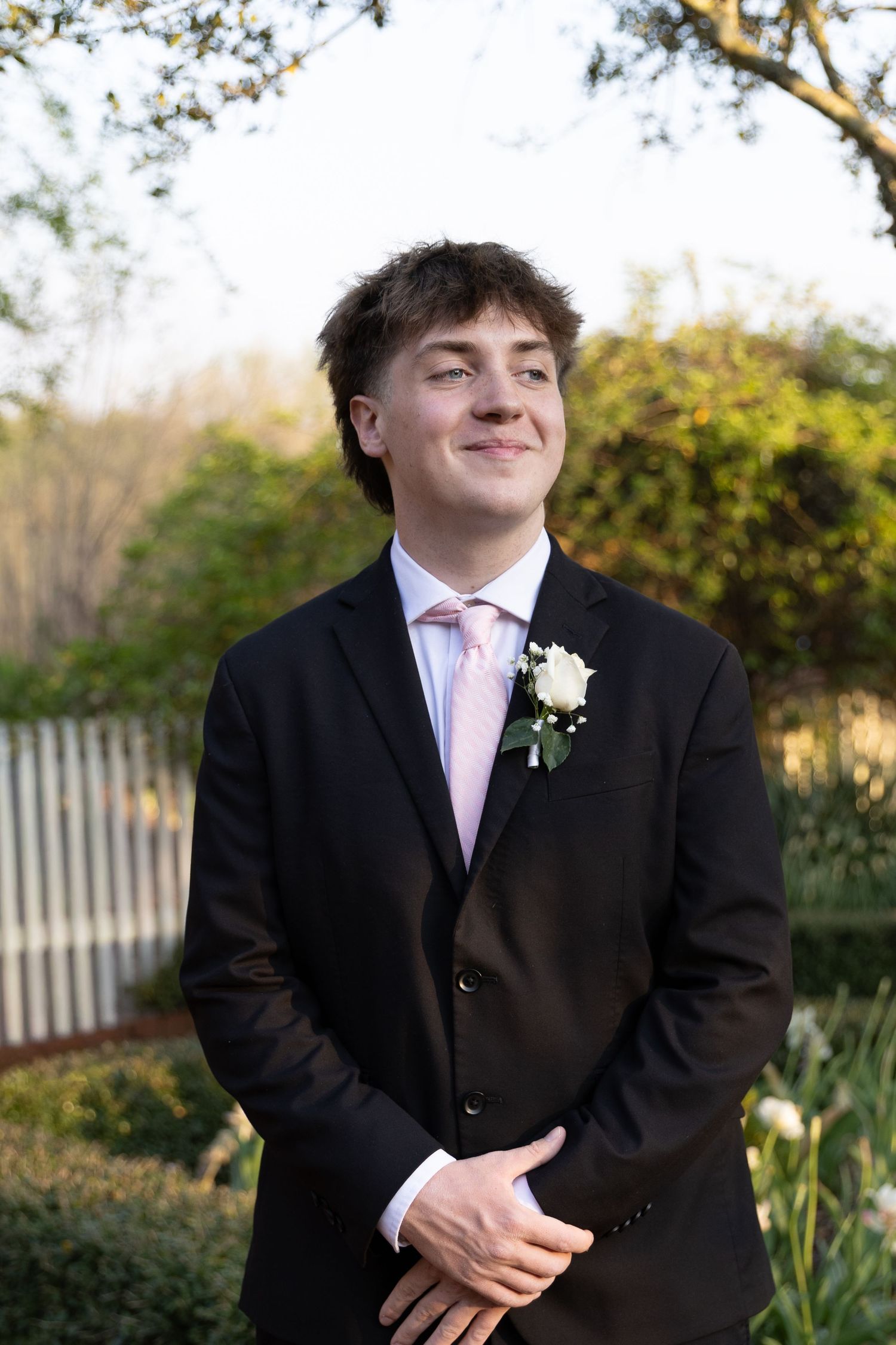 Young man in black suit with pink tie and white boutonniere smiling outdoors, dressed for prom or formal event.