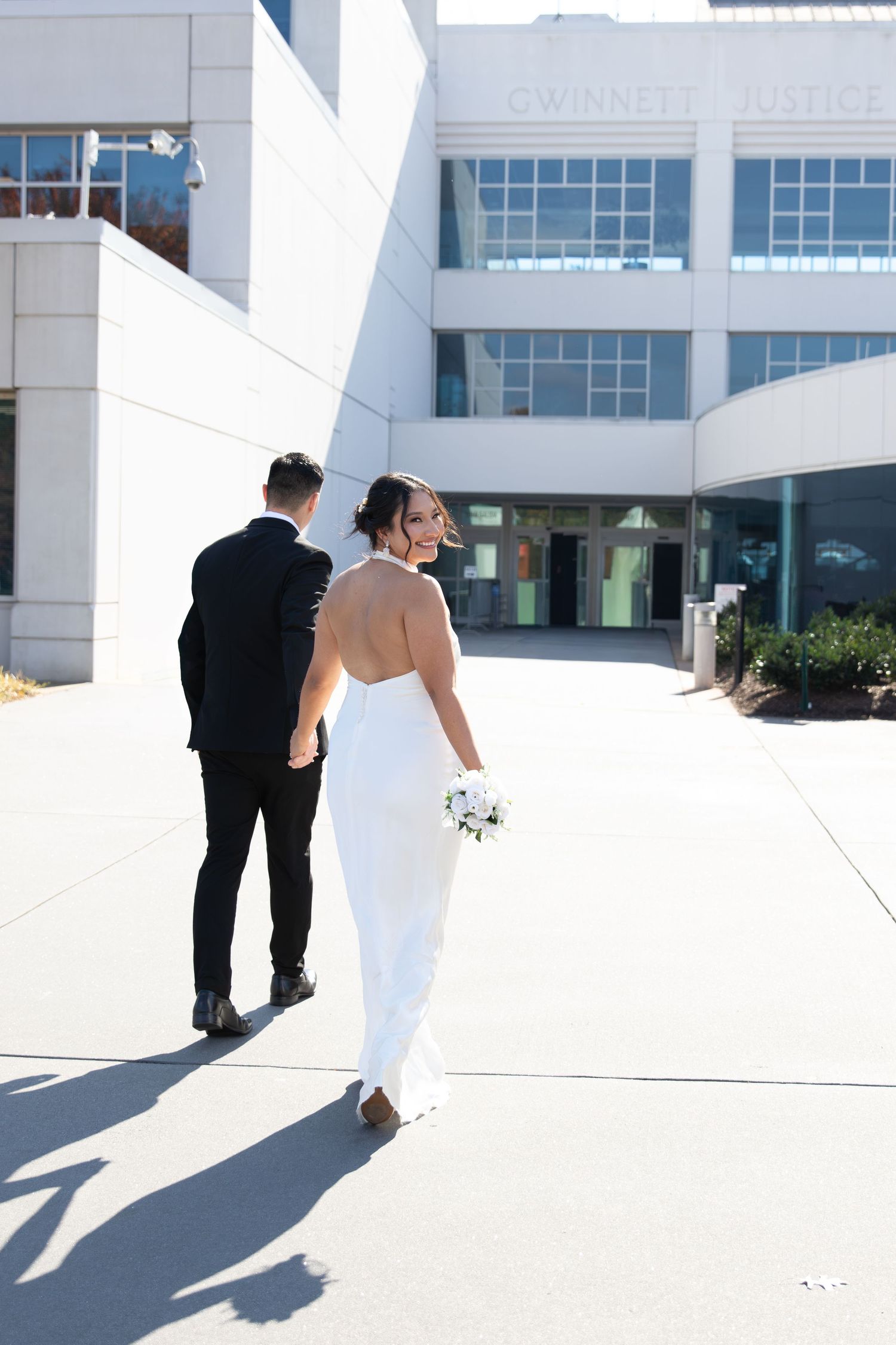 Bride in backless white gown holds bouquet walking with groom in black suit outside Gwinnett Justice Center.