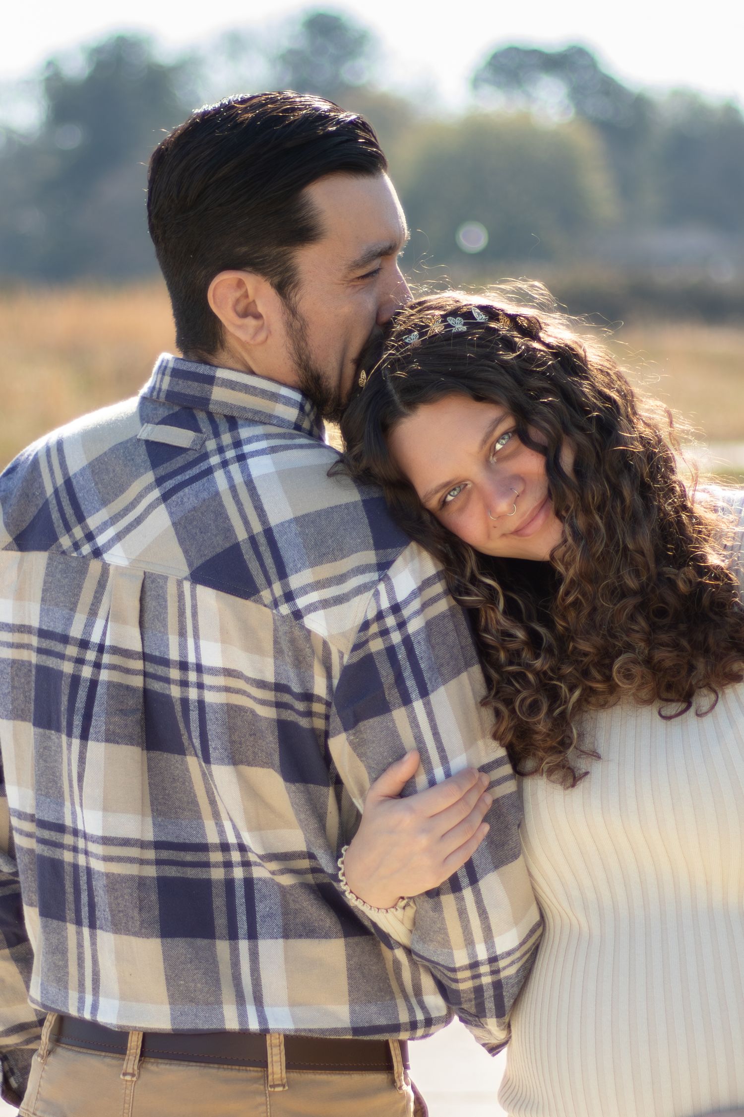 A couple embraces outdoors, man in plaid shirt kissing woman with curly hair in white sweater in a sunny field.