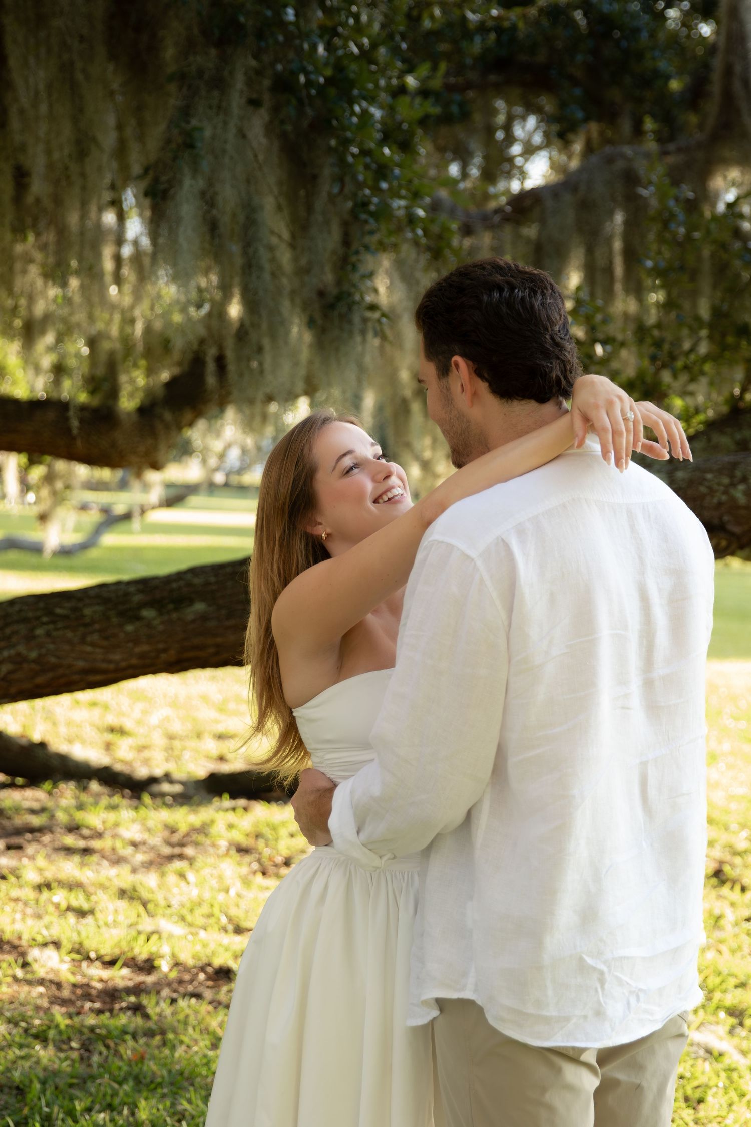 A smiling couple embraces outdoors under a moss-draped oak tree, both dressed in white attire.