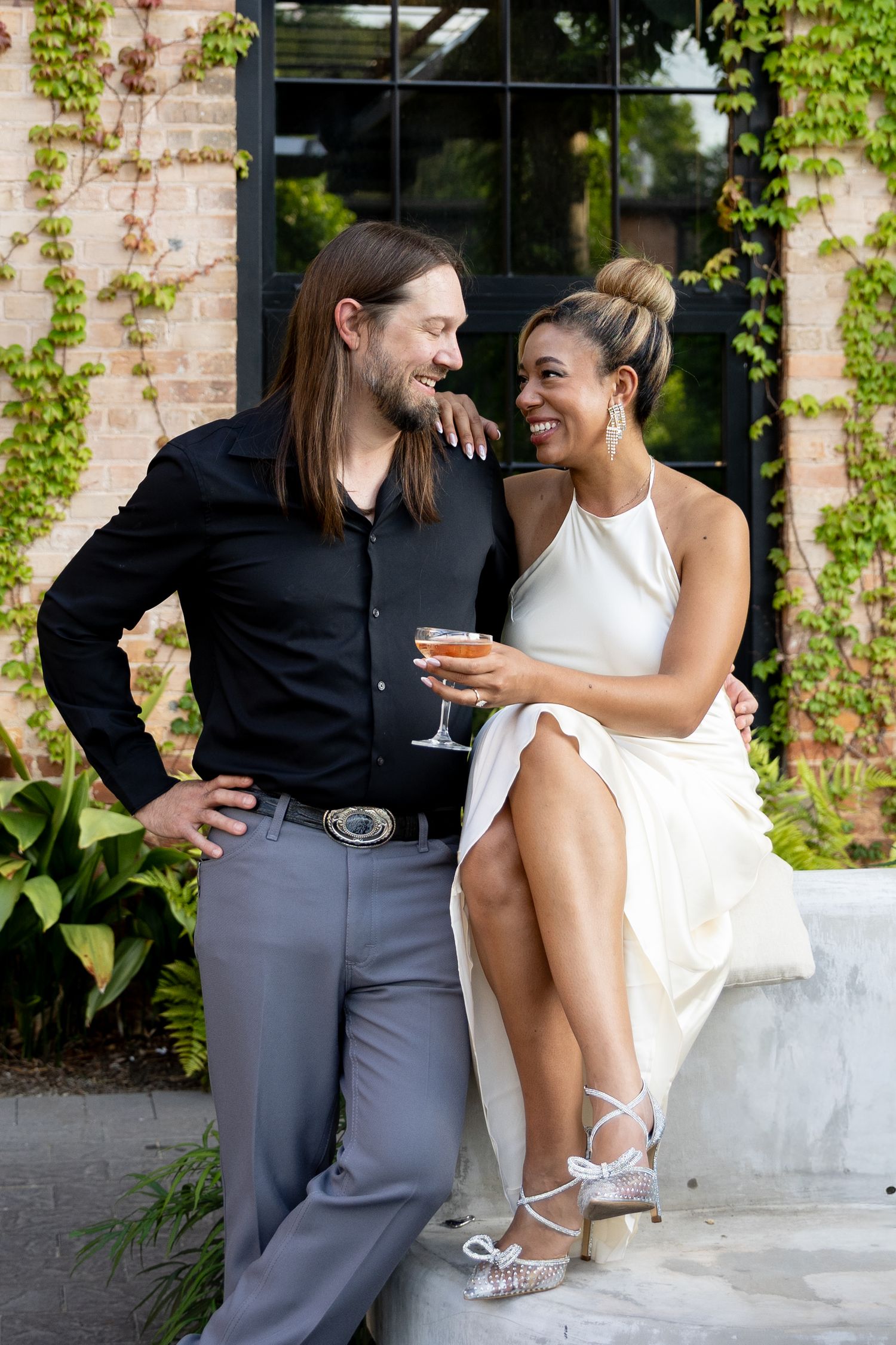 A smiling interracial couple in elegant attire share a joyful moment outdoors, woman in white dress holding champagne.