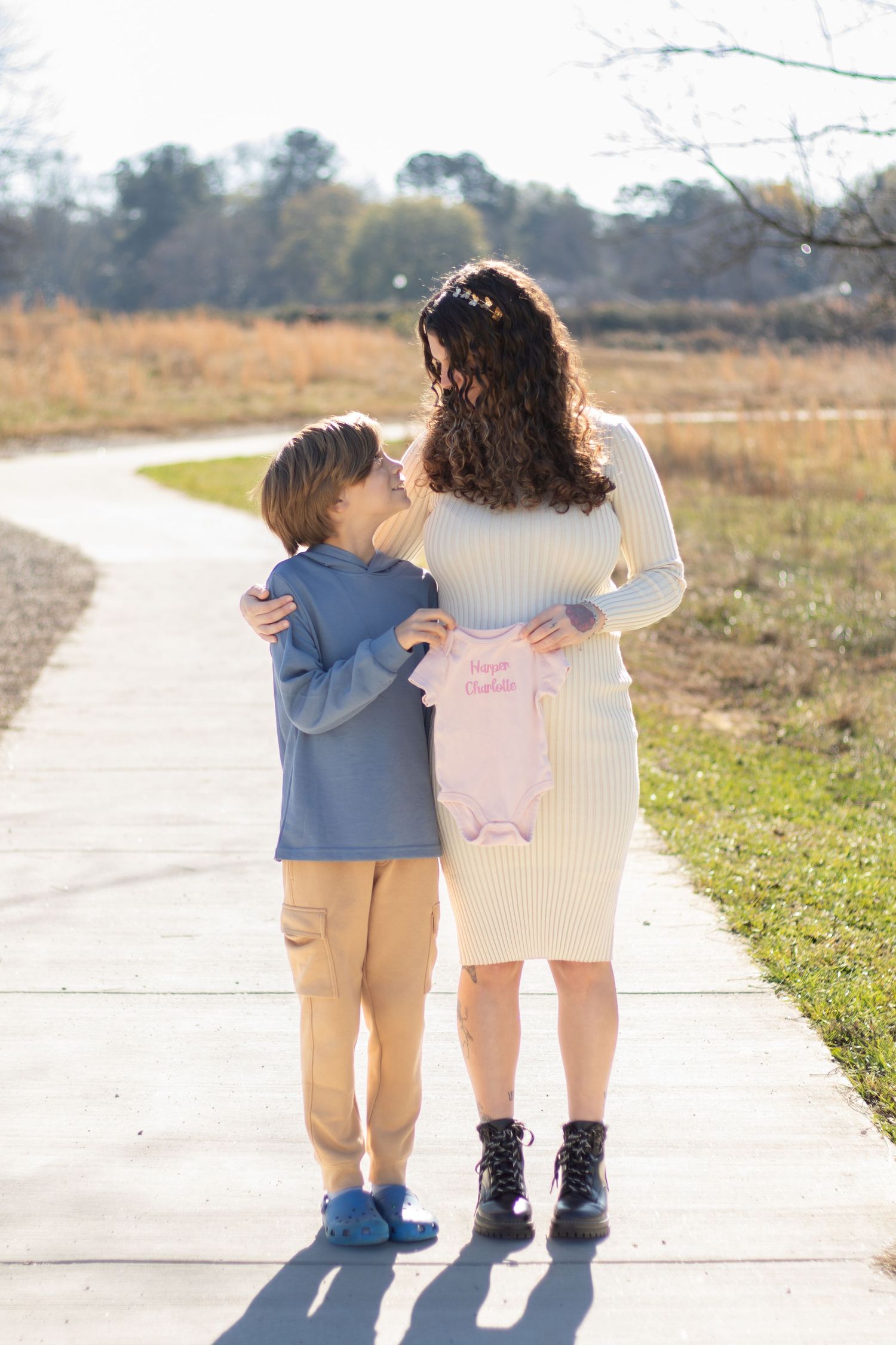 A mother and son share a warm embrace on a sunny path, holding a pink baby onesie announcing a new sibling.