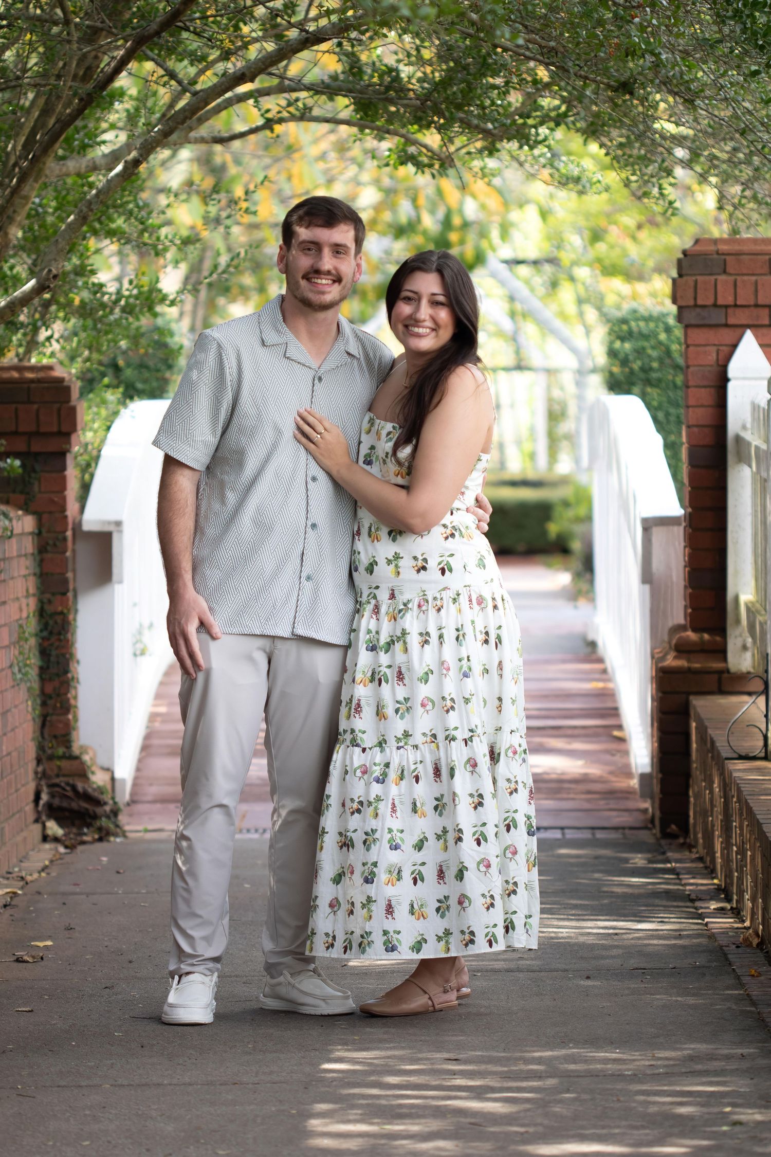 Smiling couple posing together under a green tree canopy on a brick walkway, woman in floral dress, man in gray shirt.