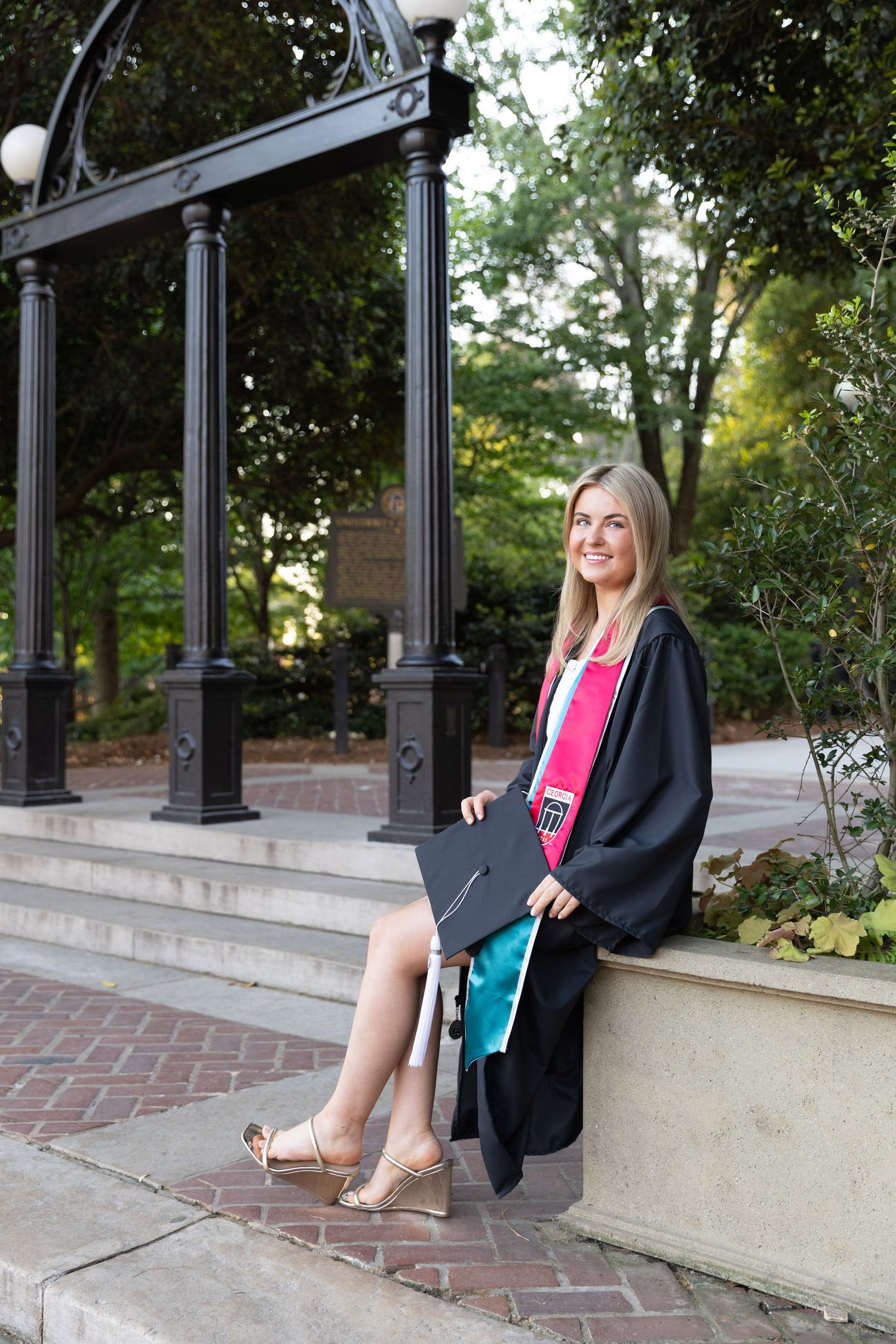 Young woman in black graduation gown with pink sash holds cap while sitting near ornate arch outdoors.