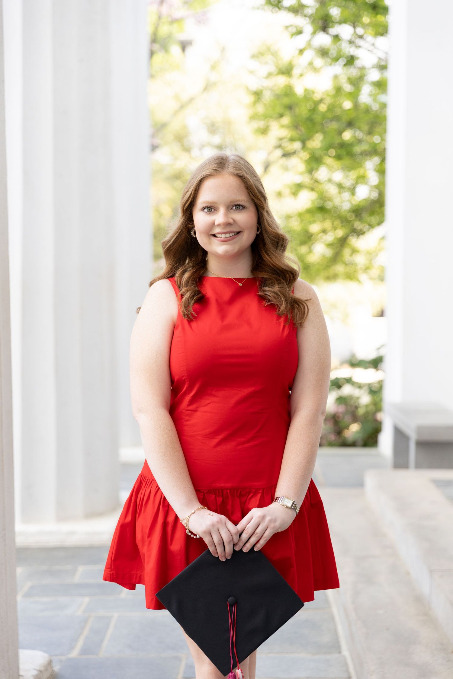 Young woman in red dress holding graduation cap smiles outdoors near white columns.