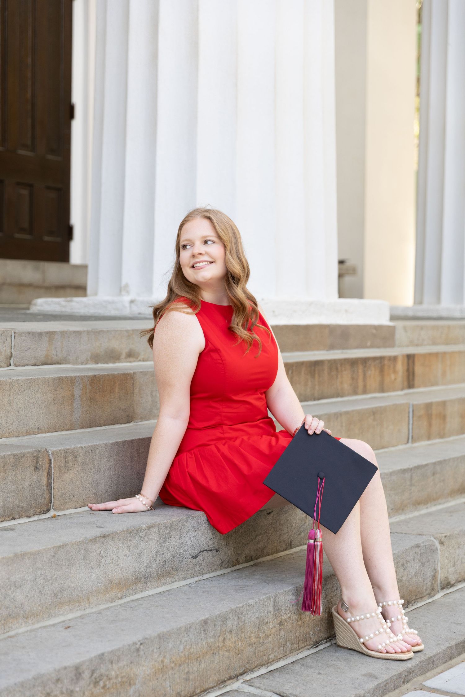 Young woman in red dress holding graduation cap sits on stone steps, smiling, with white columns behind her.