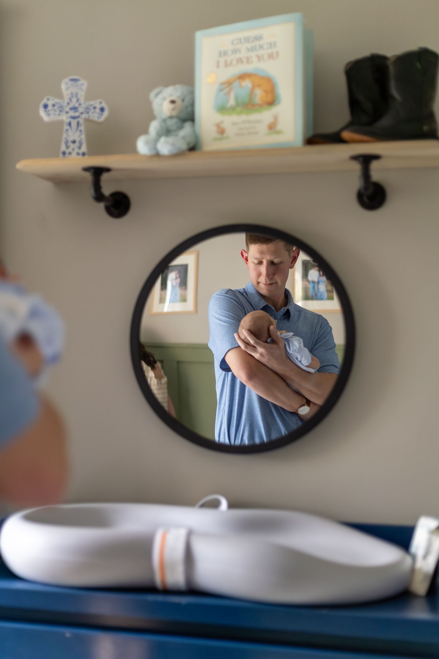 Father cradling newborn baby reflected in round nursery mirror above changing table with shelf decor.
