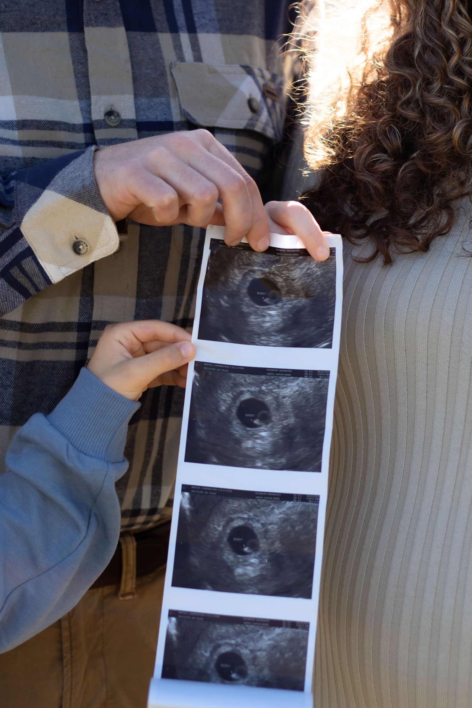 Couple holding ultrasound sonogram strip showing baby scans, pregnancy announcement photo outdoors.