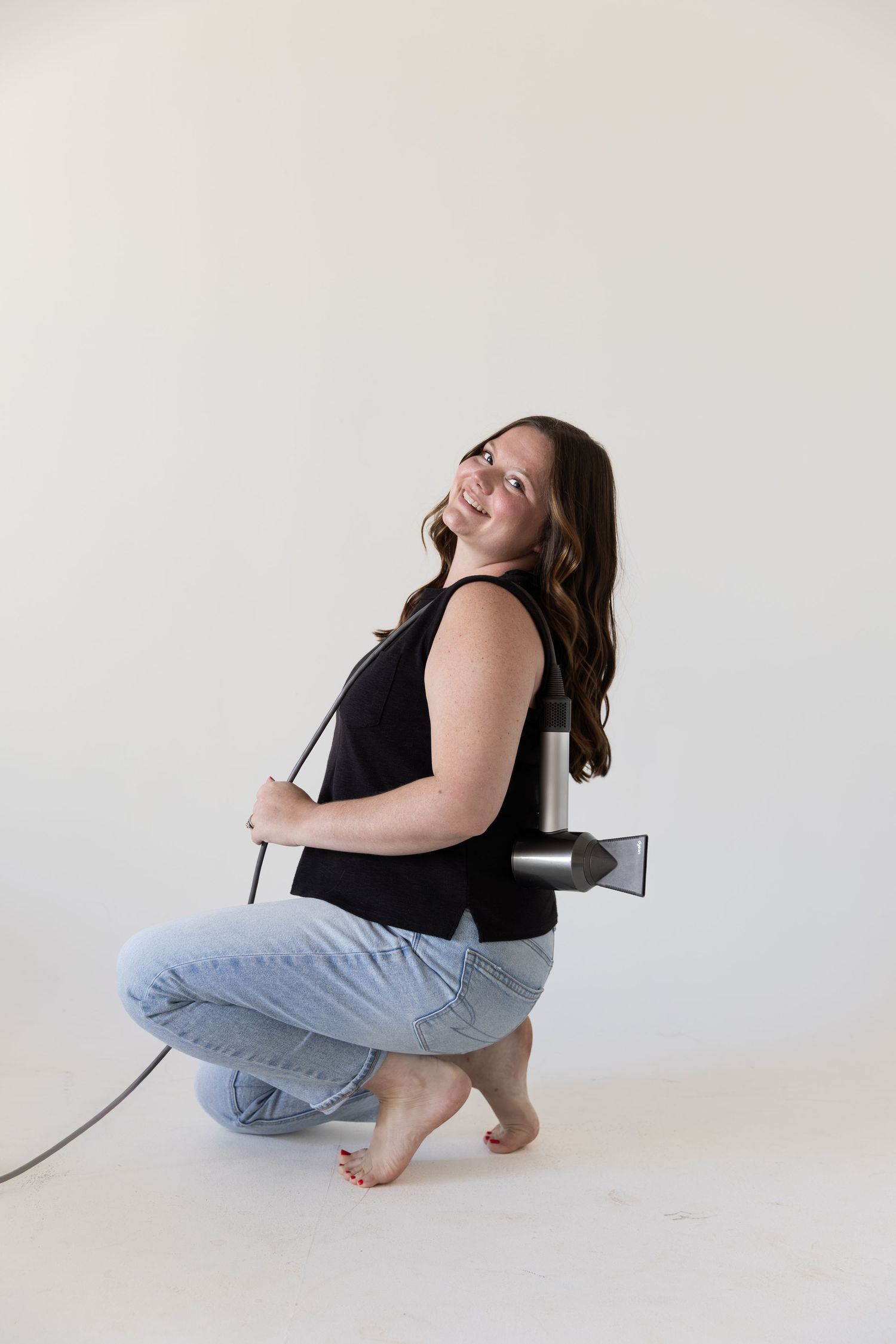 Smiling woman crouching barefoot in jeans and black top, holding a hair dryer against white background.