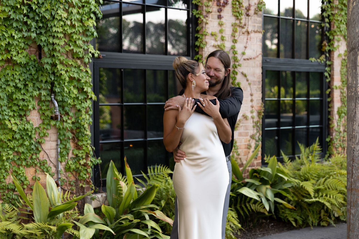 Couple embracing outdoors in front of ivy-covered brick wall, woman in white satin slip dress, man in dark suit.