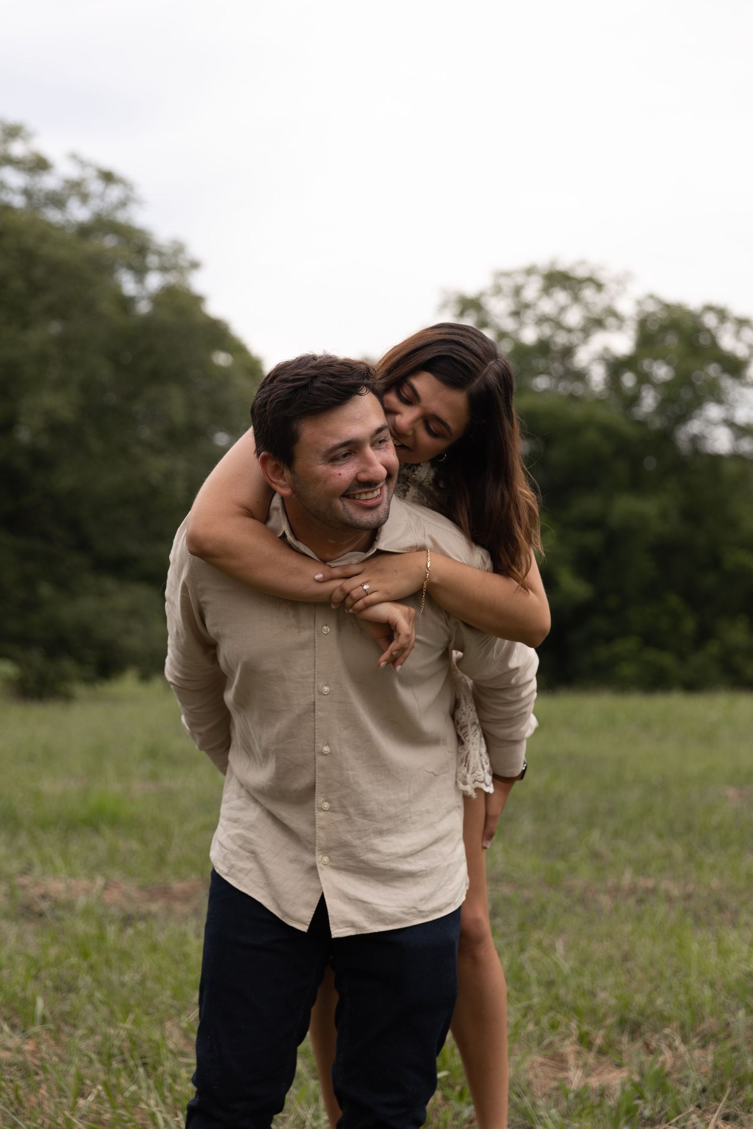 Smiling groom giving his bride a piggyback ride in a green field during romantic engagement photos.