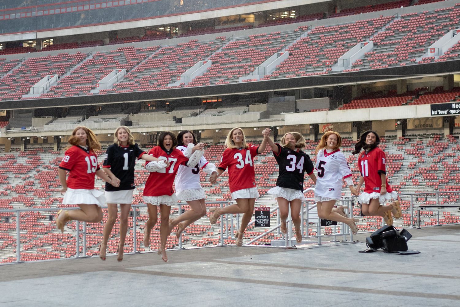 Group of women in football jerseys jumping in unison at a large empty stadium on a sunny day.