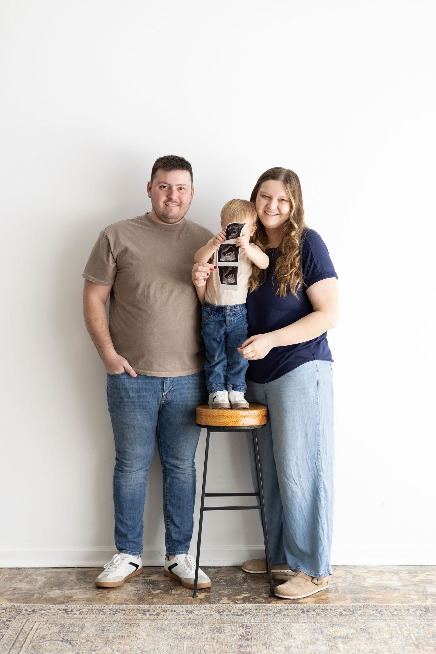 A family of three poses against a white wall, parents flanking a toddler on a stool holding ultrasound photos.