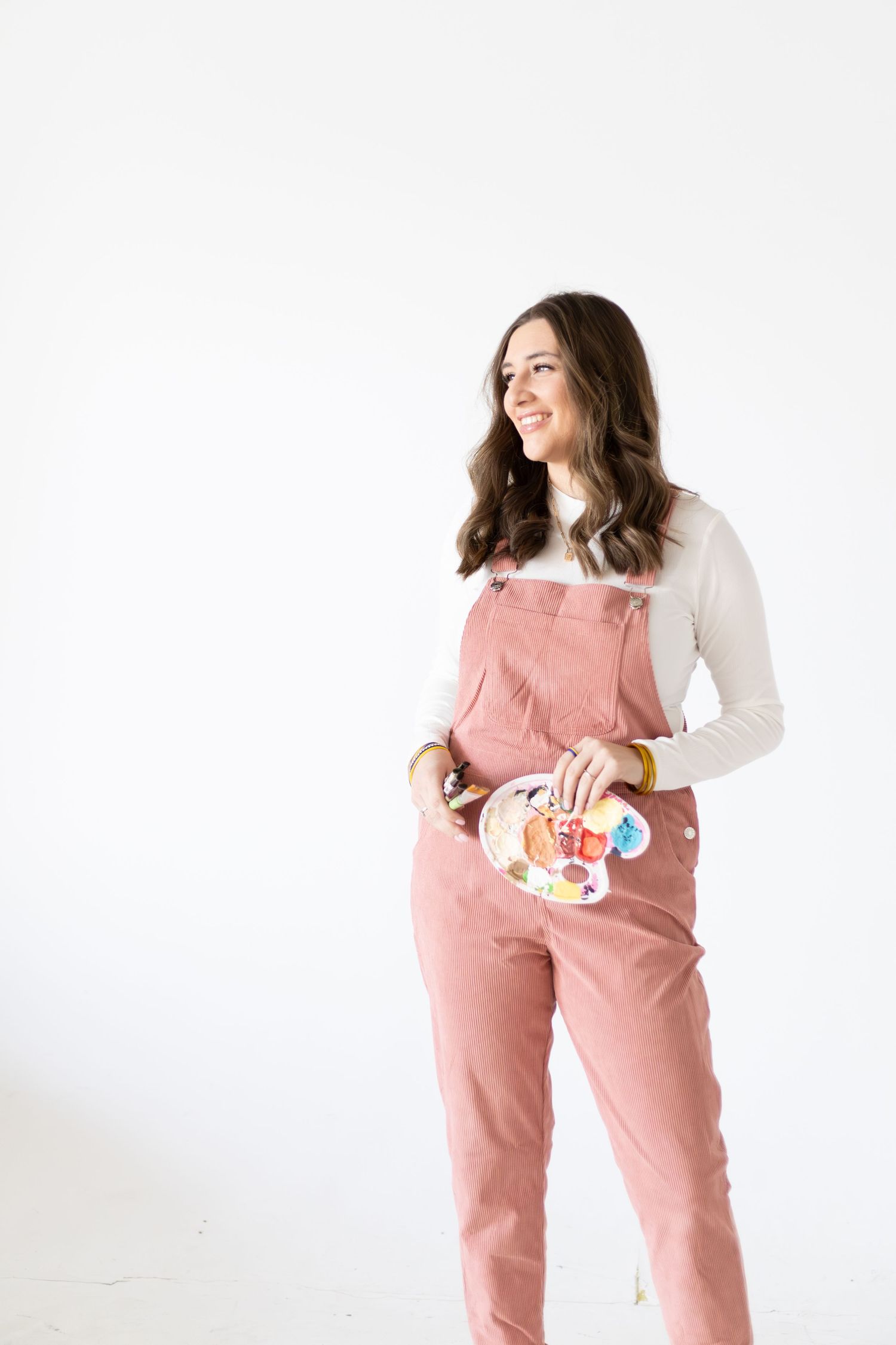 Smiling woman in pink overalls holding a paint palette, standing against a white background.