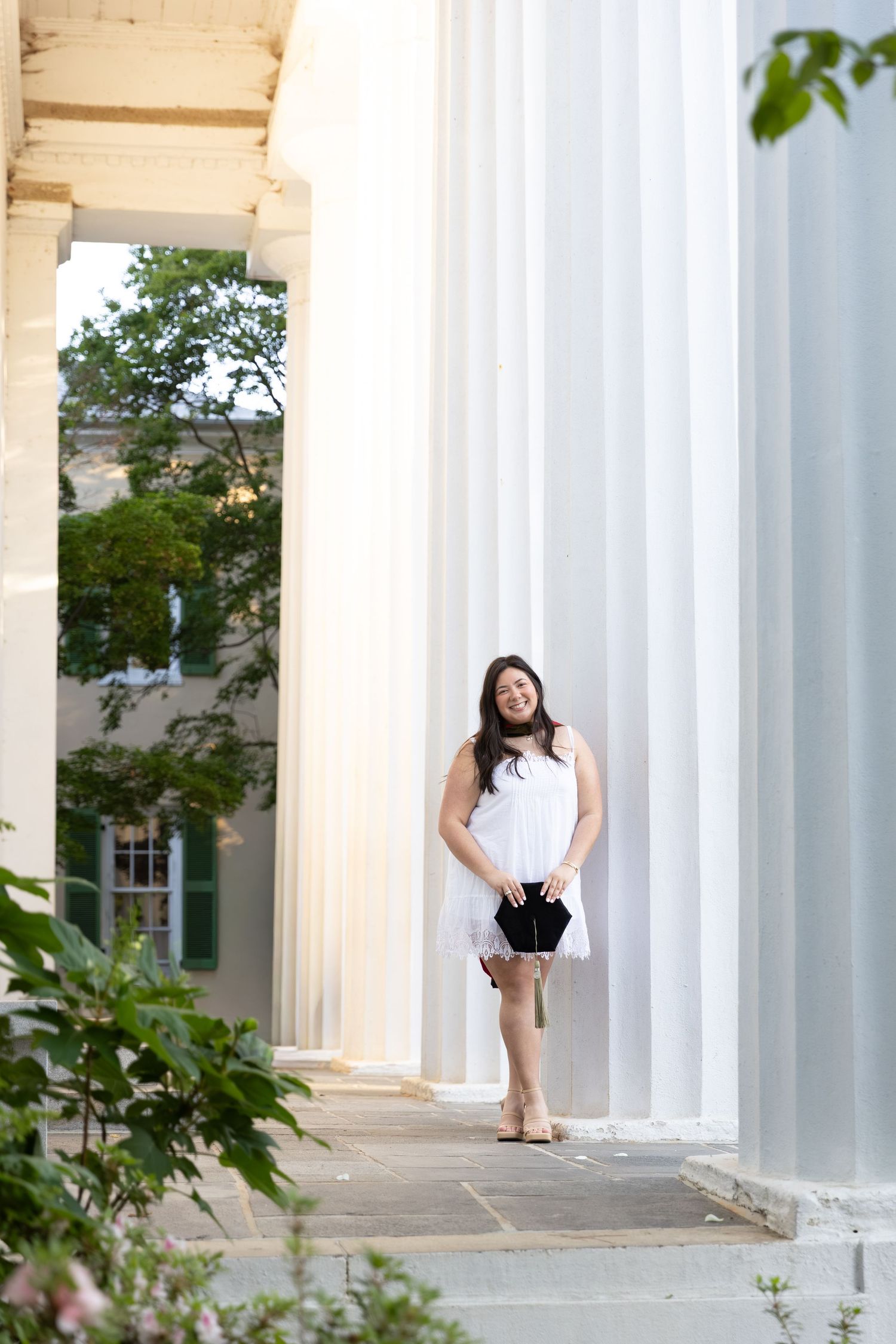 Young woman in white dress posing between large white columns of a classical building surrounded by green trees.