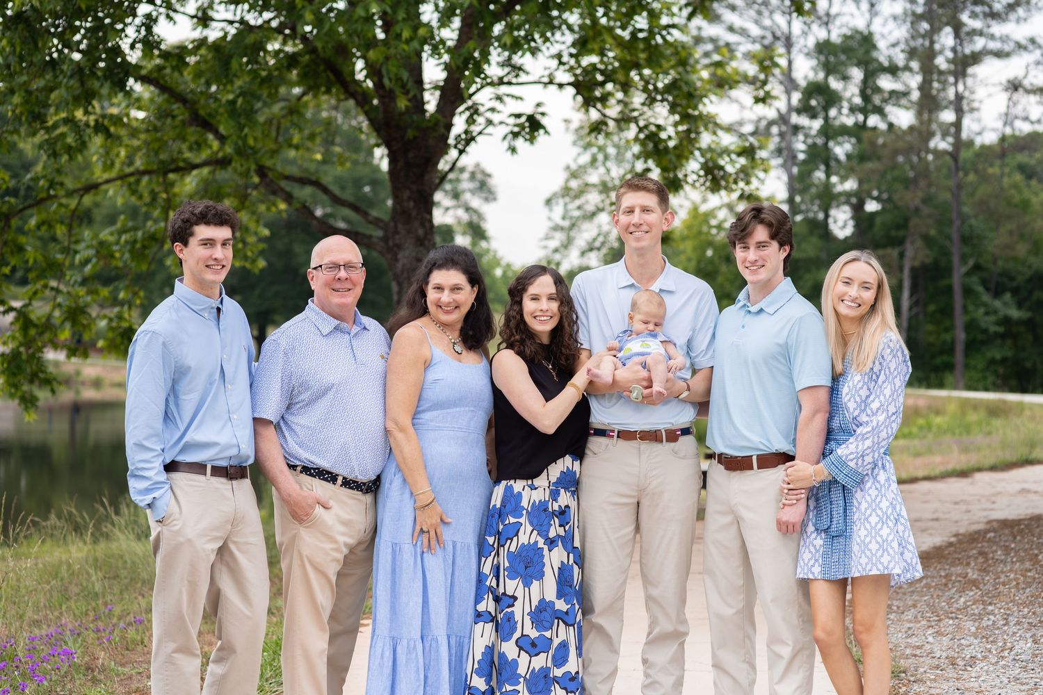 A joyful family of seven poses outdoors near trees, dressed in coordinating blue and khaki outfits, with one member holding a baby.