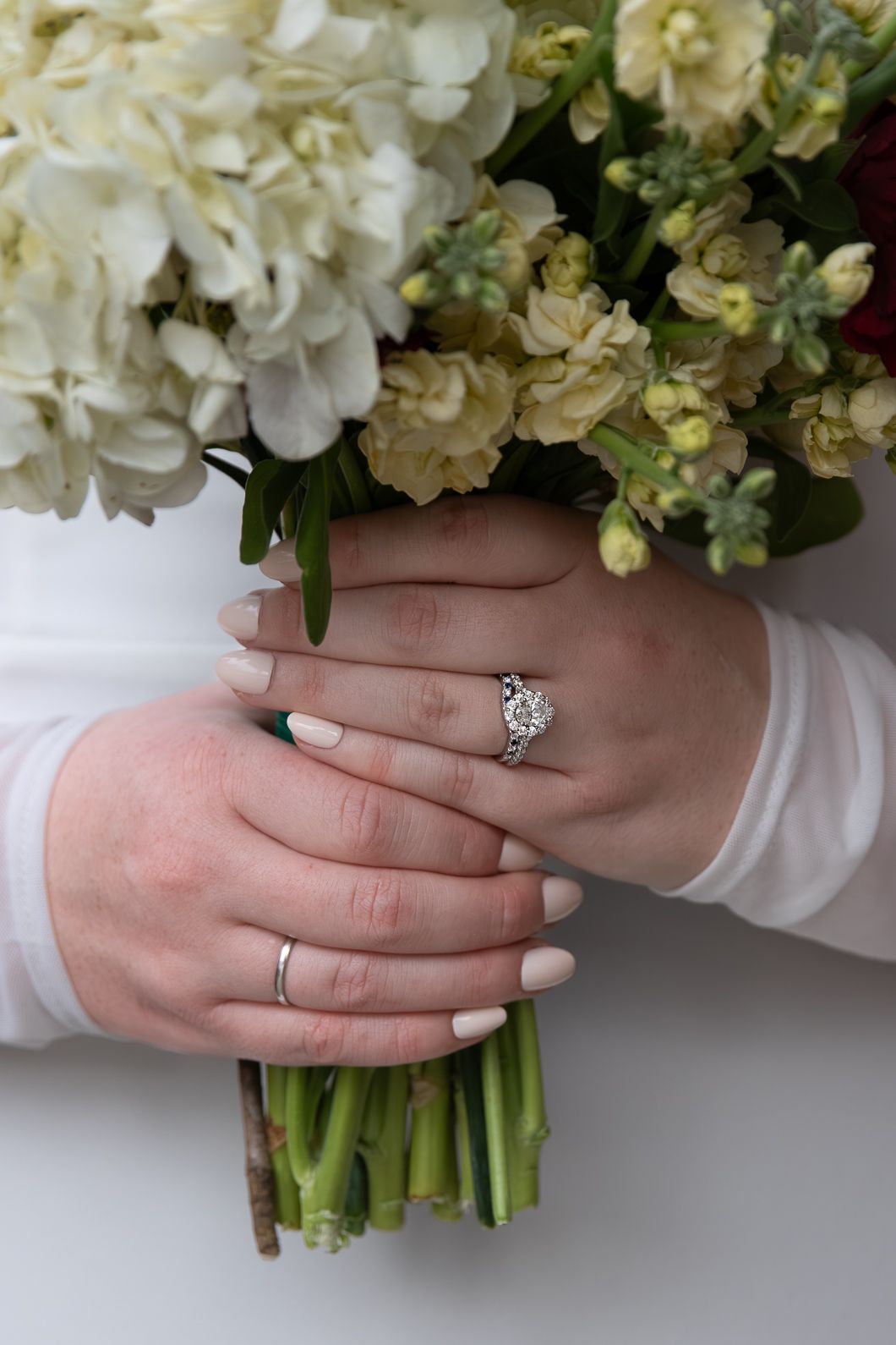 Close-up of bride's hands holding white floral bouquet, wearing diamond engagement ring and wedding band.