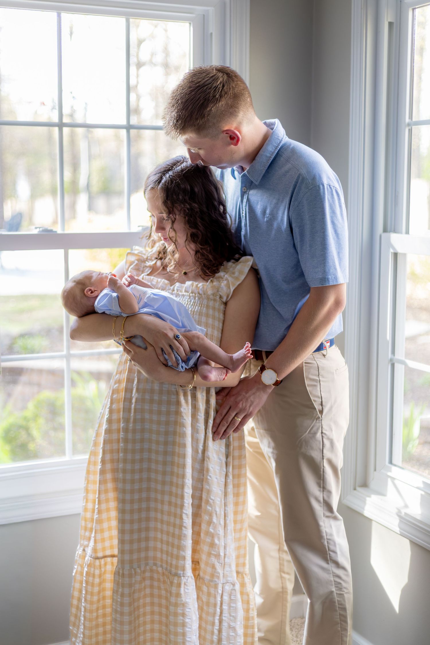 New parents sharing a tender moment with their newborn baby by a bright window, mother in yellow gingham dress.