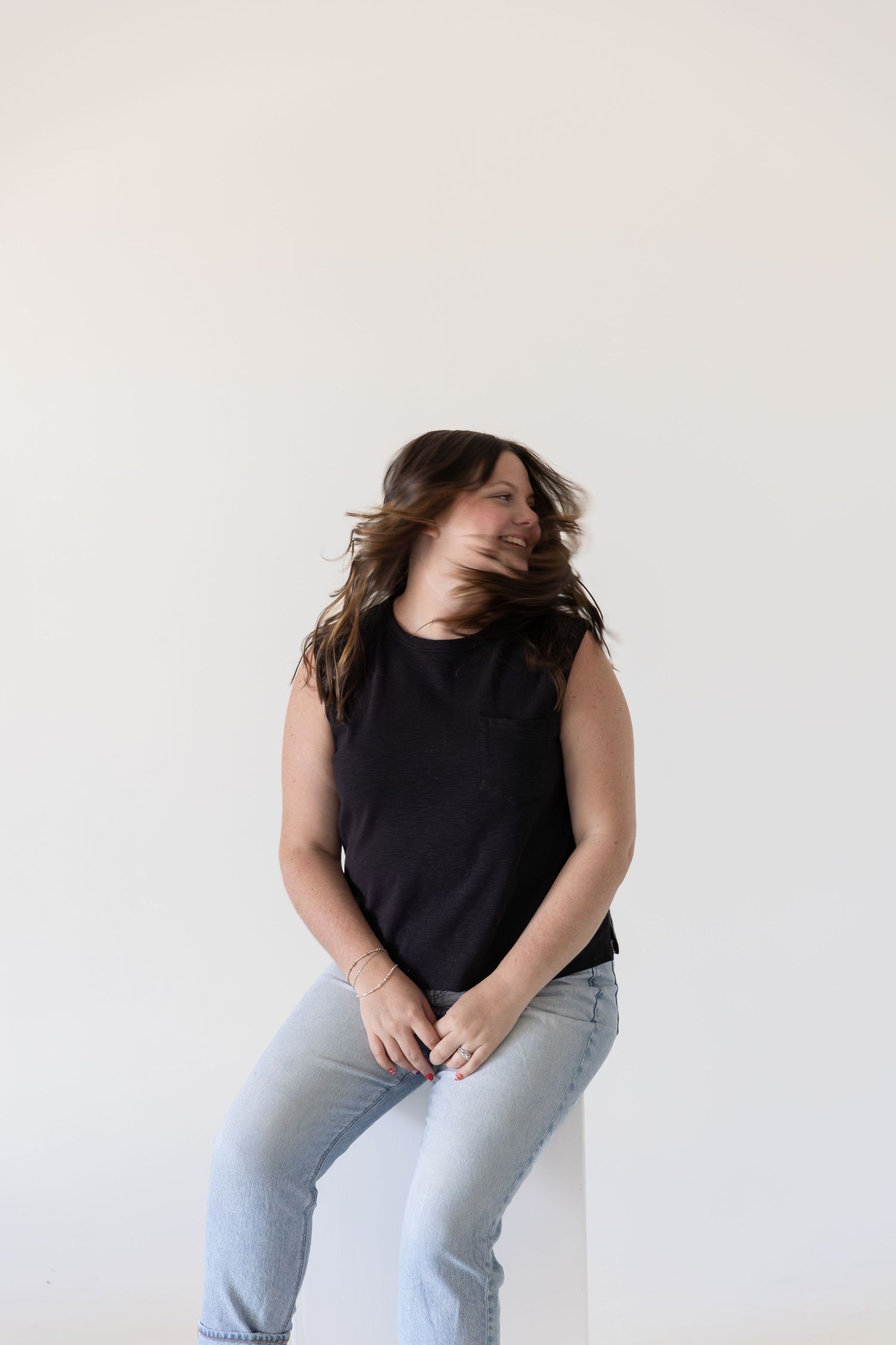 Woman in black sleeveless top and light jeans posing with hair tossing motion against white background.