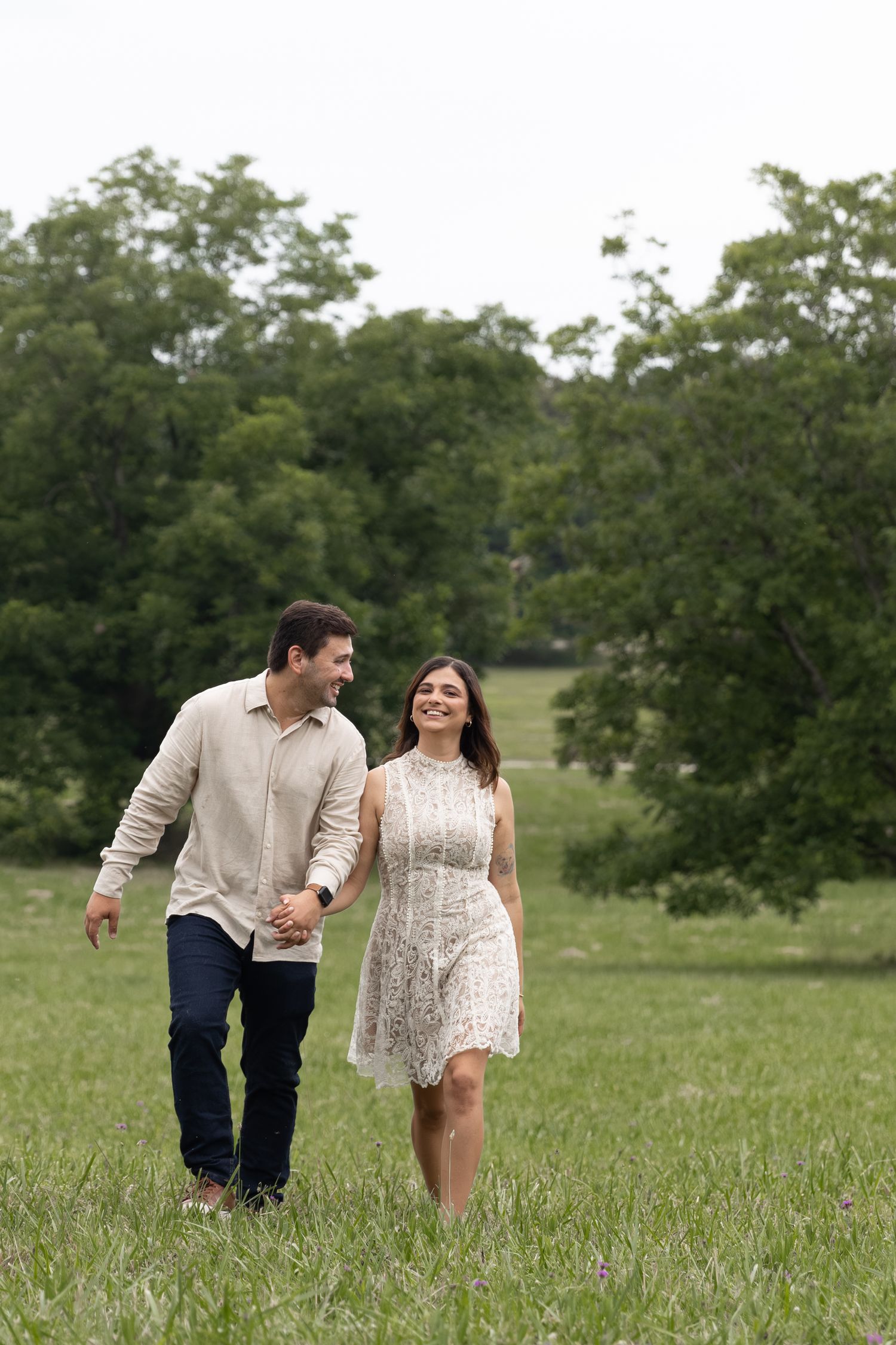 A happy couple holds hands while walking through a lush green field, smiling at each other on a cloudy day.