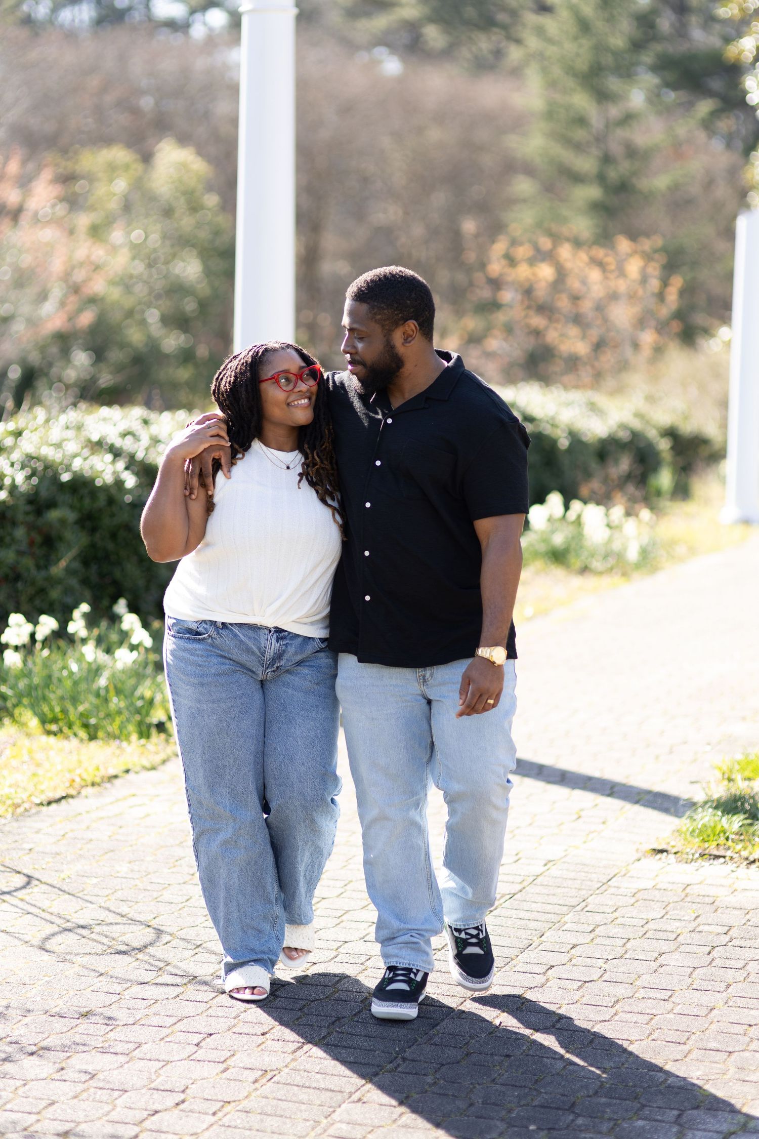 A couple walks arm-in-arm on a sunny brick path, smiling at each other in a garden setting.