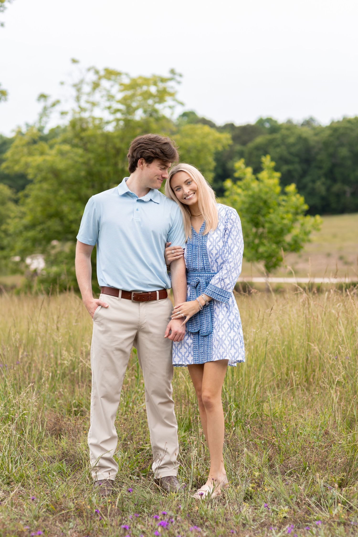 Young couple poses in a field, man in blue polo and khakis with woman in blue patterned dress, surrounded by greenery.