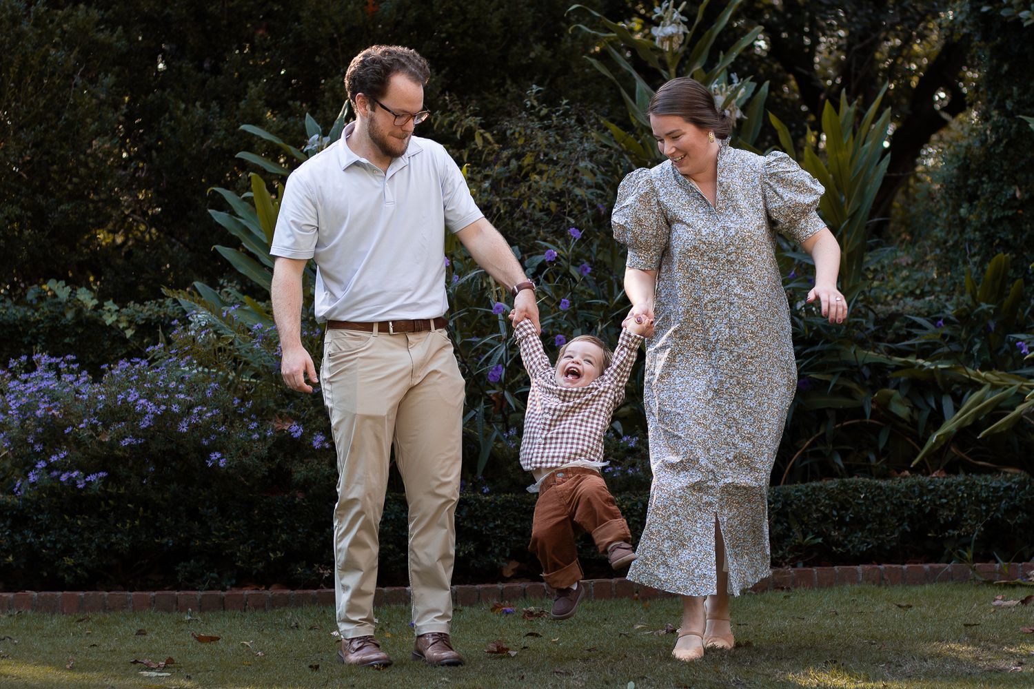 A happy family with parents swinging their toddler by the hands in a lush green garden.