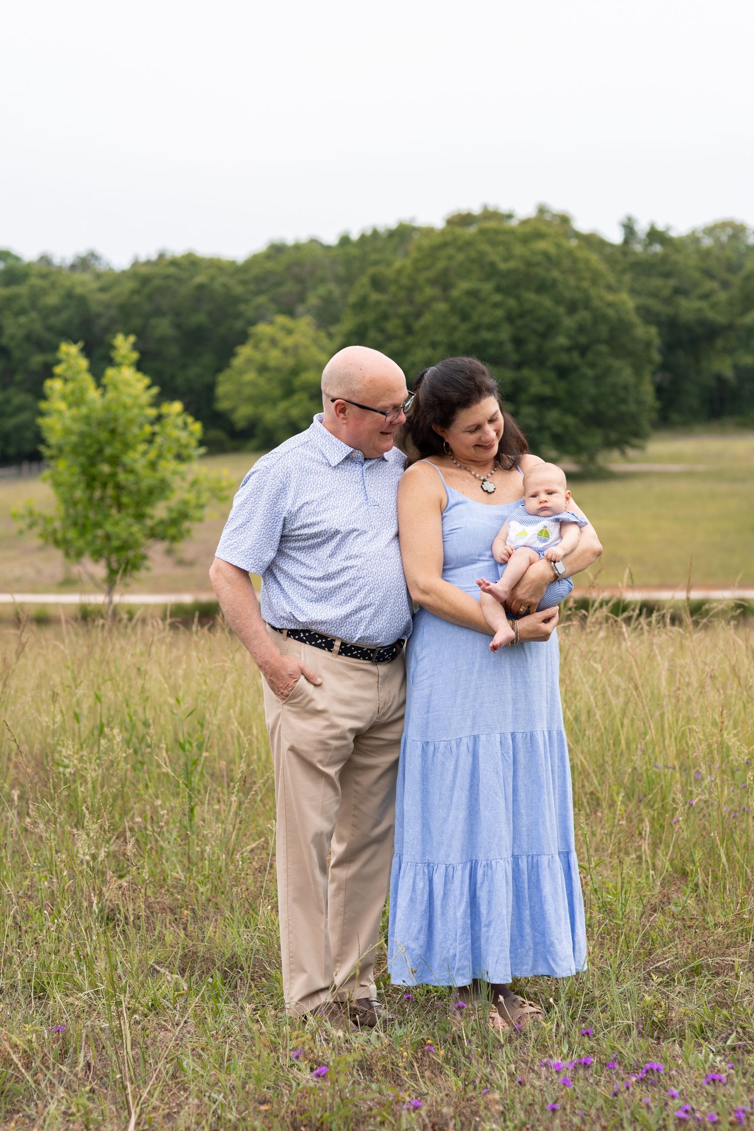 Grandparents smiling at baby in a sunny field, woman in blue dress holding infant, man in light blue shirt beside her.