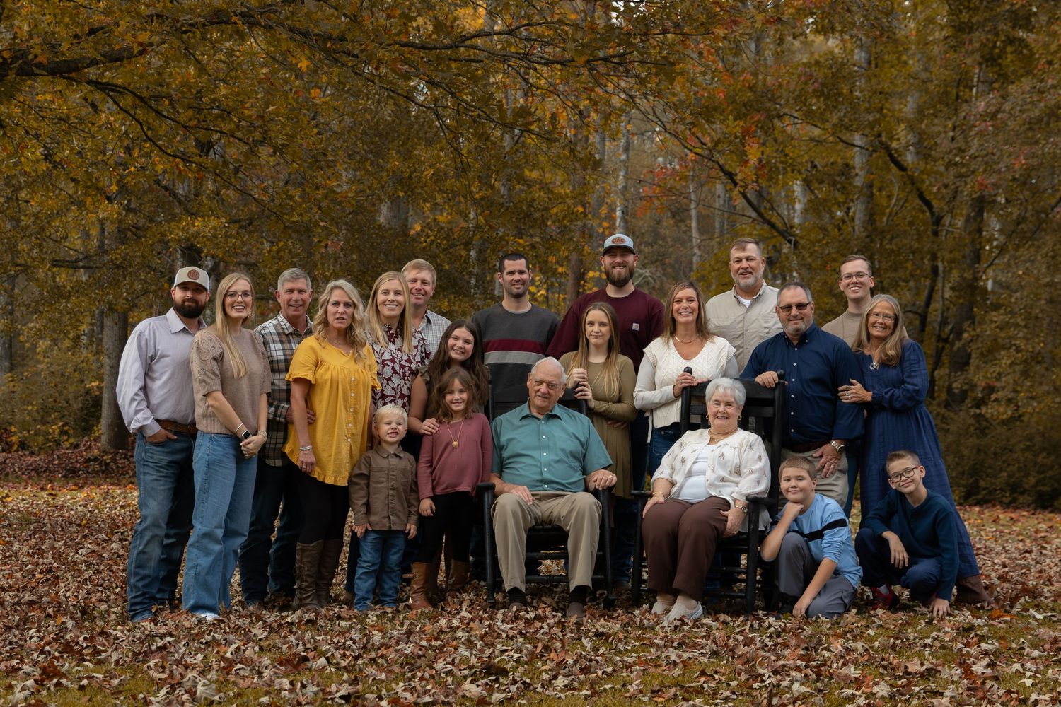 Large multigenerational family portrait outdoors in autumn, posing among colorful fall foliage and leaves on the ground.