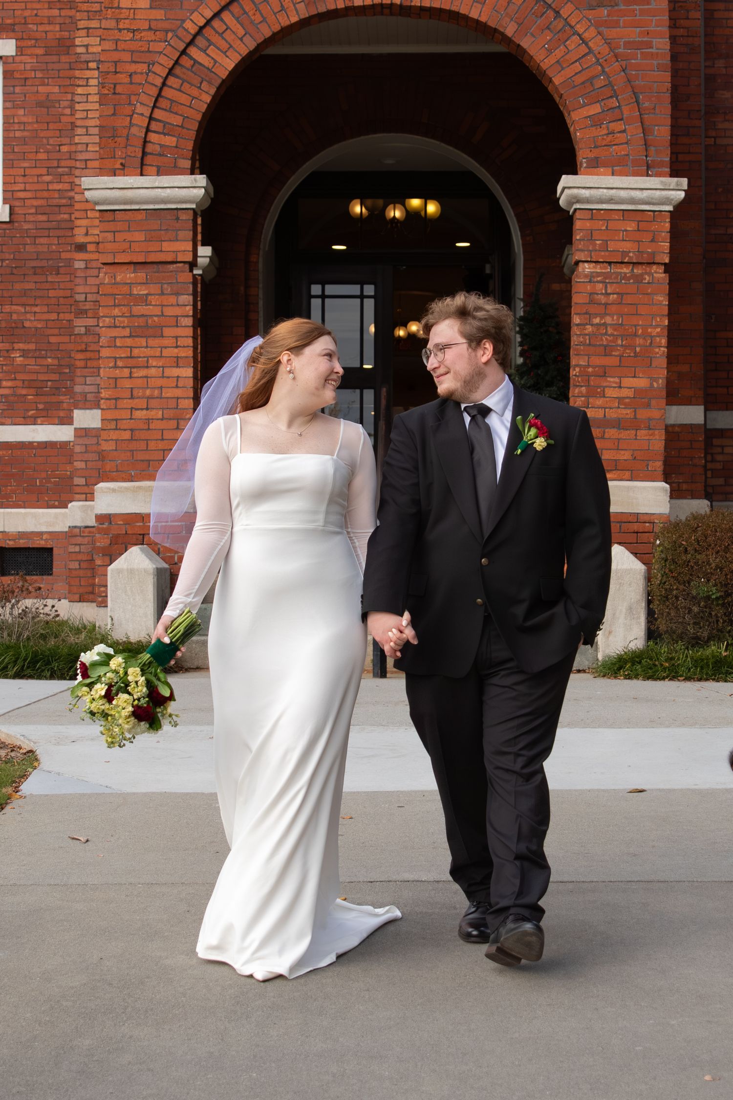 A bride and groom walk hand-in-hand outside a brick church, smiling at each other on their wedding day.