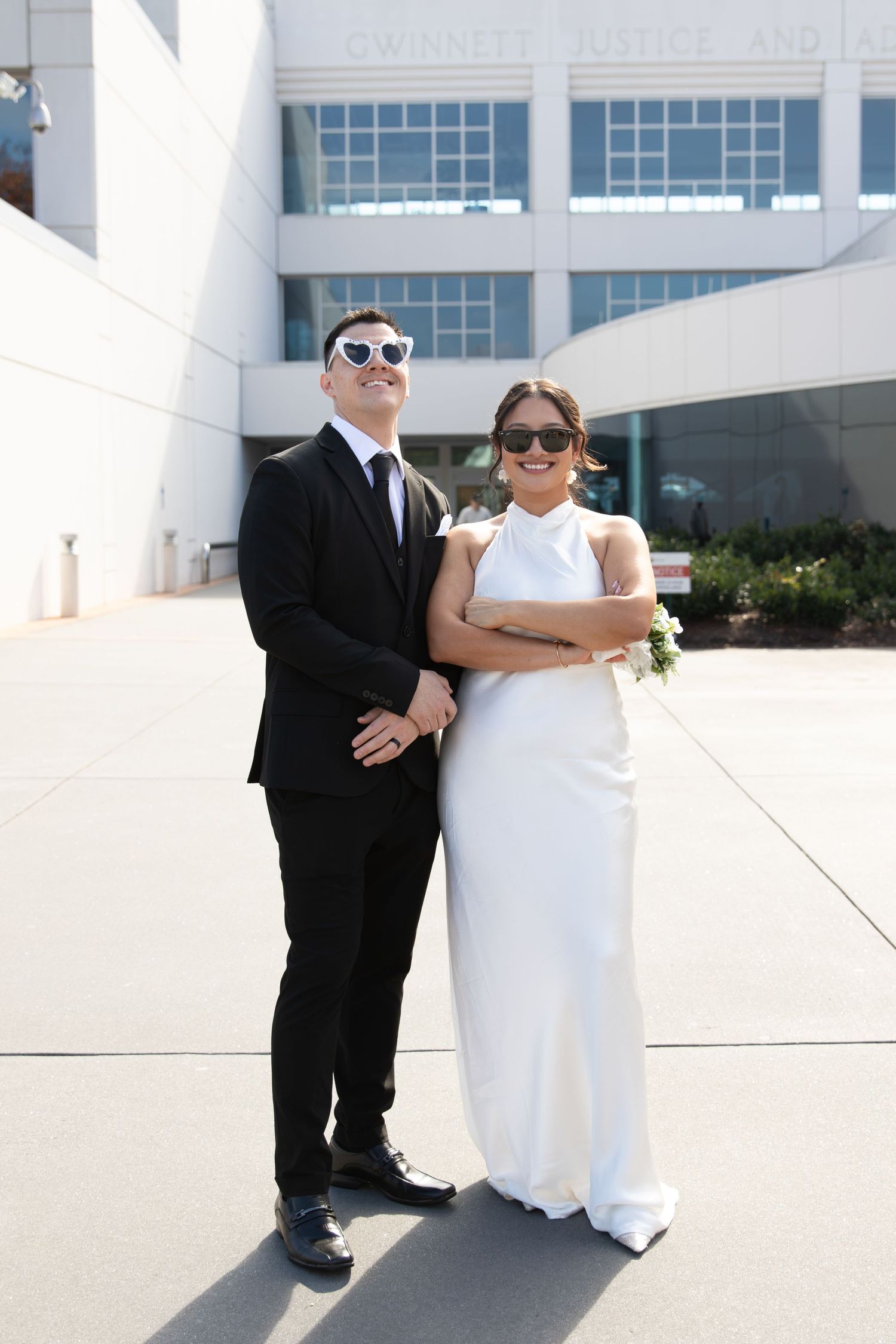 Stylish newlyweds wearing sunglasses outside Gwinnett Justice Center, groom in black suit, bride in white fitted gown.