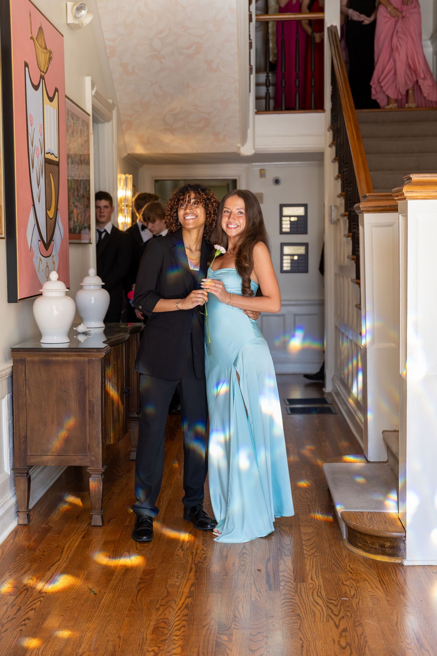 Teenage couple dressed in prom attire pose in elegant home hallway, girl in blue gown, boy in black tuxedo.