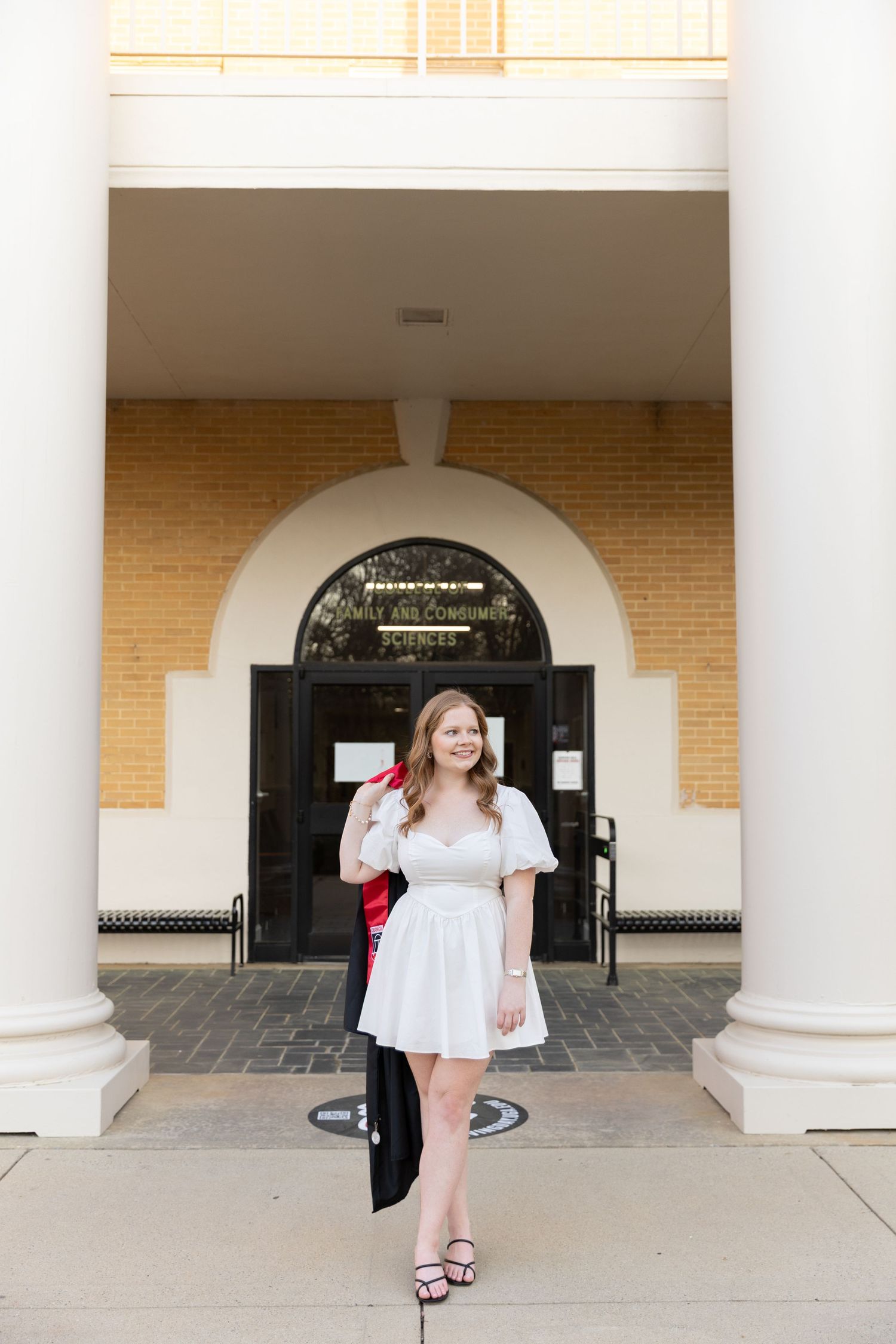 Young woman in white dress holding graduation regalia outside a university building entrance.