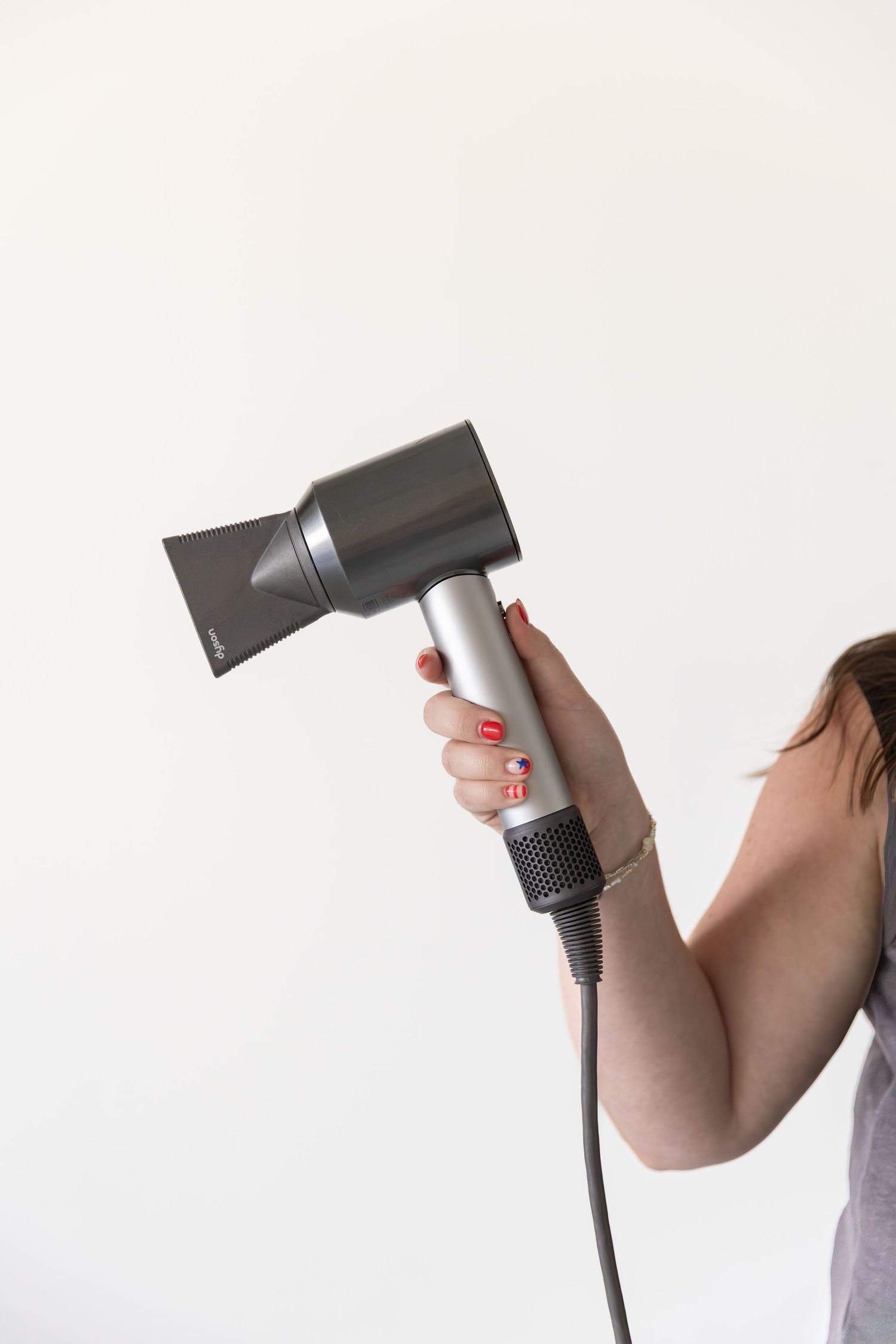 Woman holding a modern Dyson hair dryer with red manicured nails against a white background.