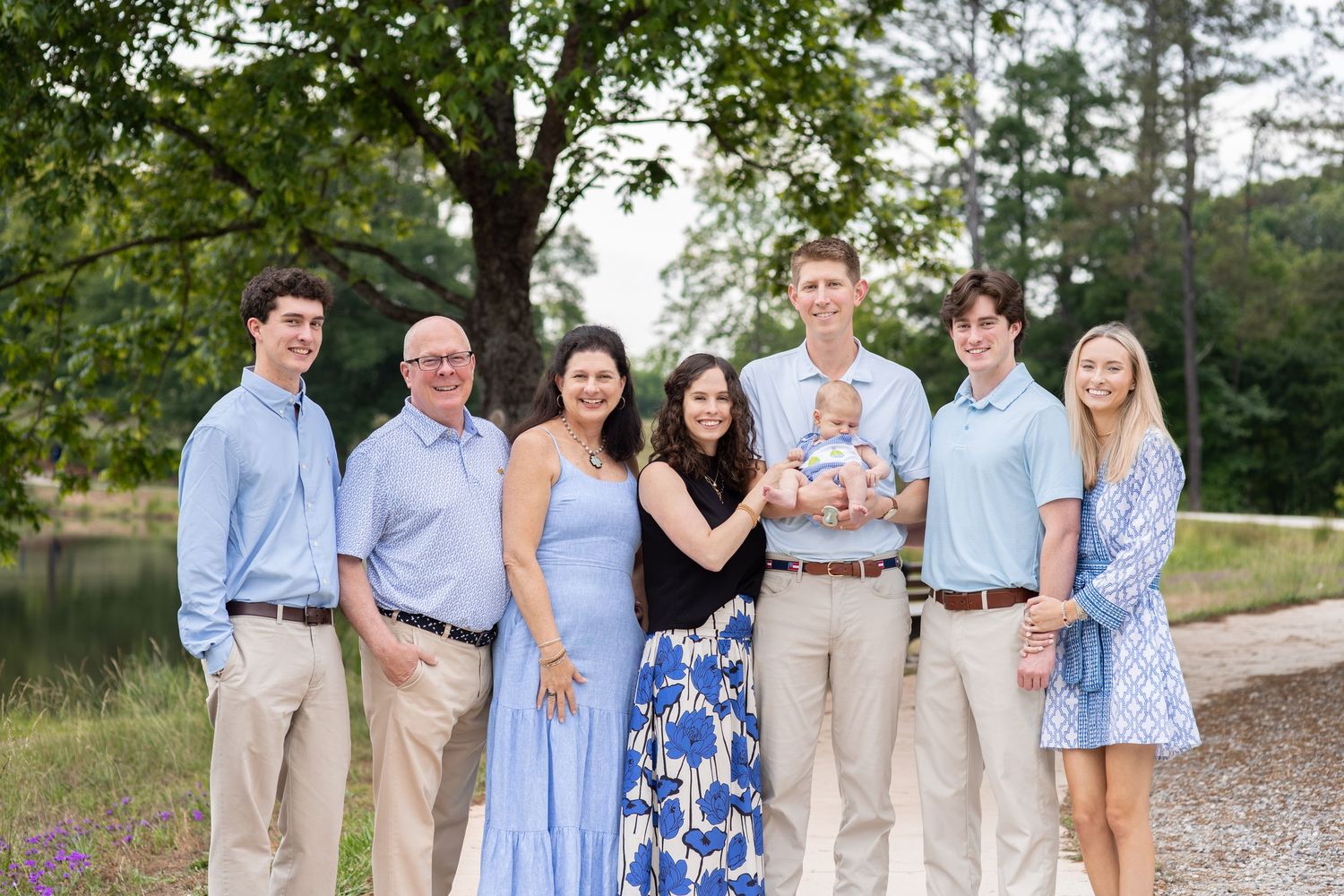 A joyful family of seven poses outdoors near a tree, dressed in blue and white outfits, with one member holding a baby.