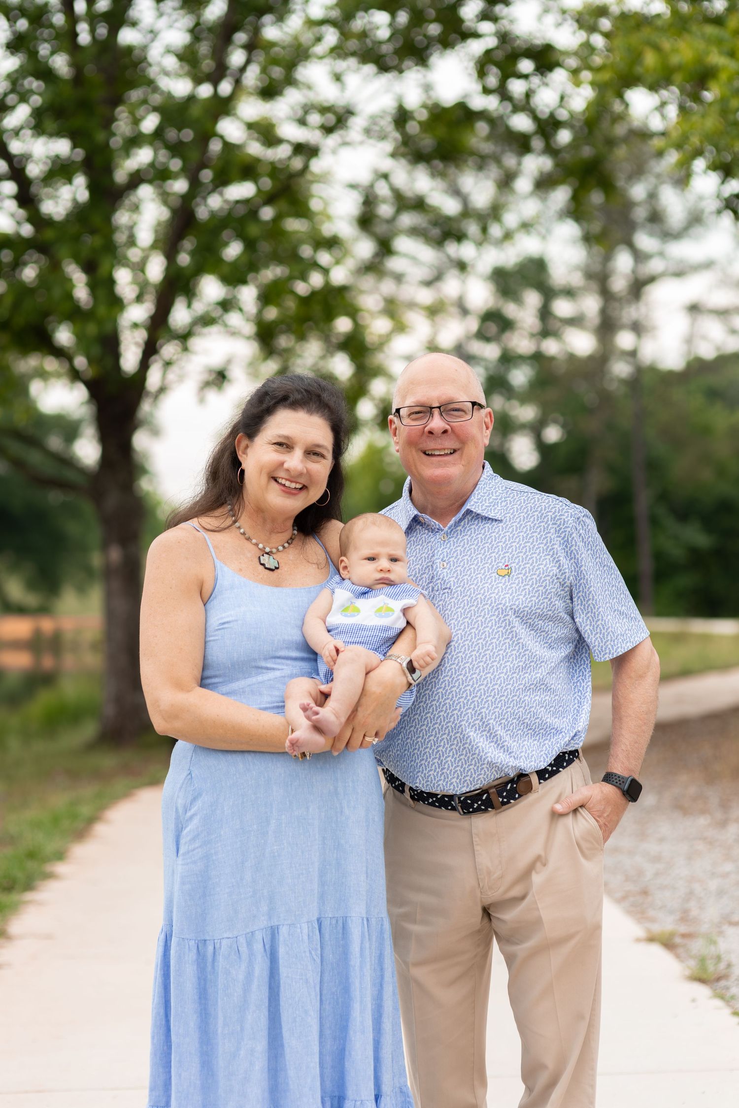 Smiling grandparents holding a baby outdoors on a sunny day, dressed in blue, with lush green trees in the background.