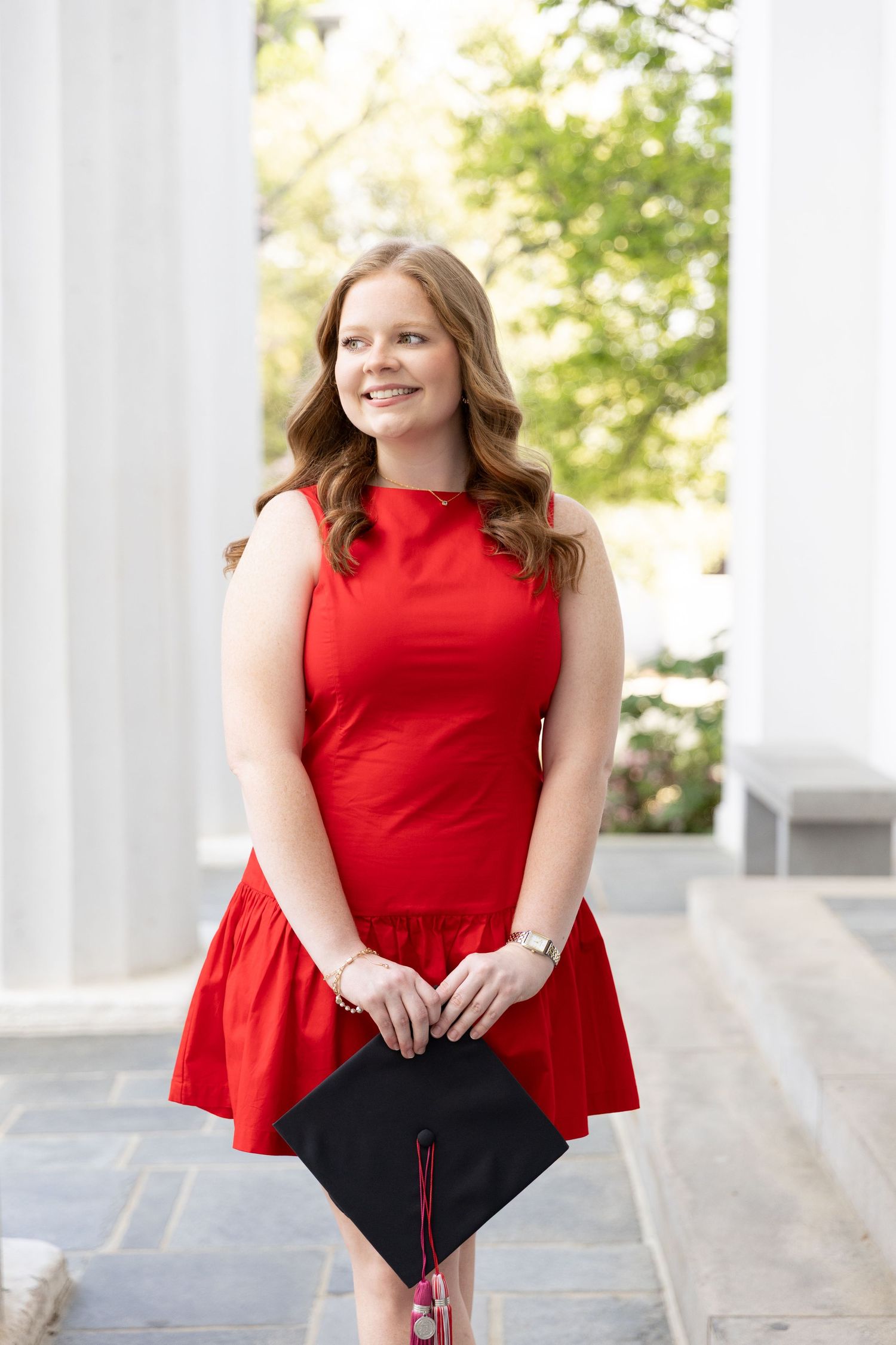 Smiling young woman in red dress holding graduation cap between white columns outdoors.