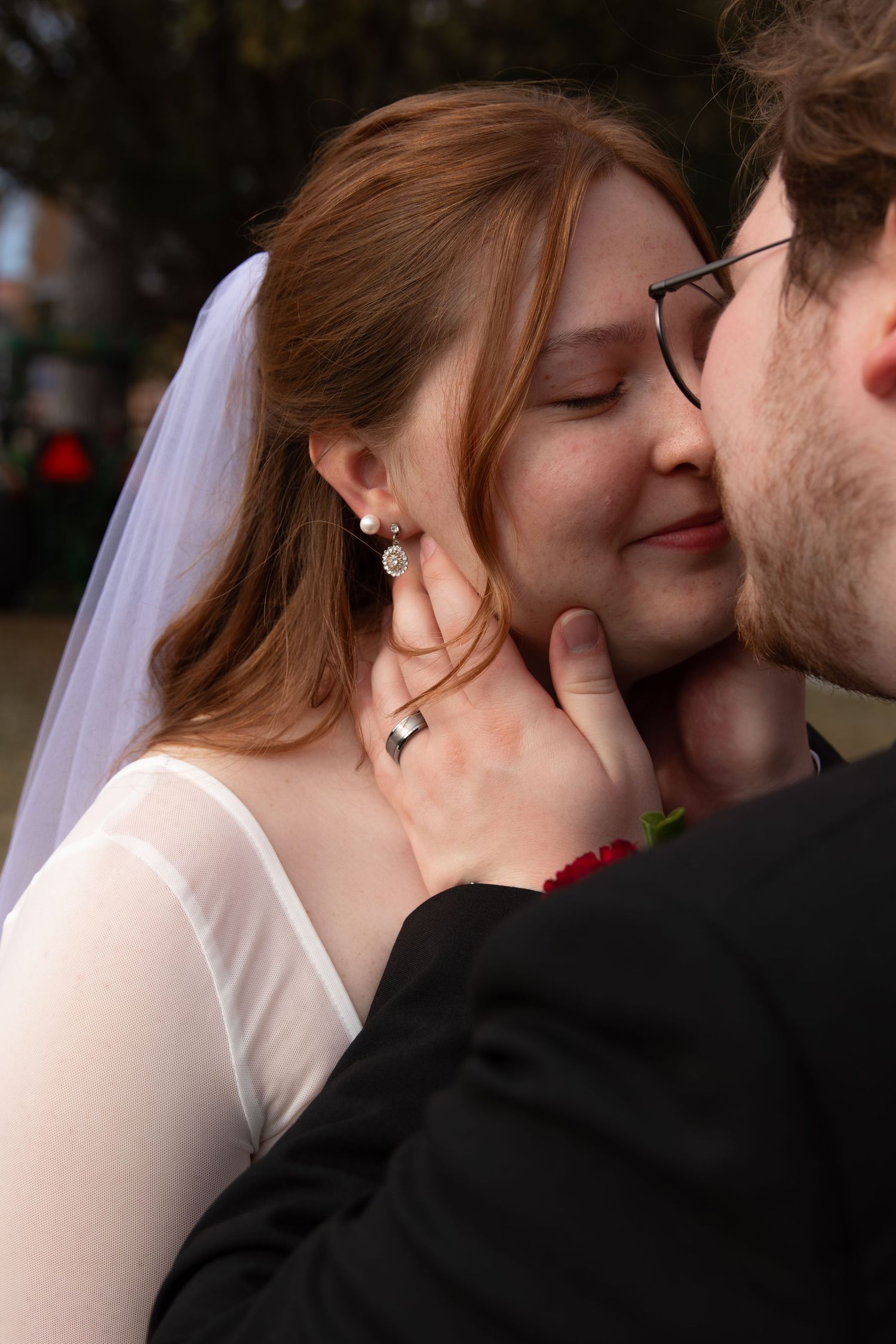 Close-up of smiling bride in white veil being kissed by groom in black suit outdoors.