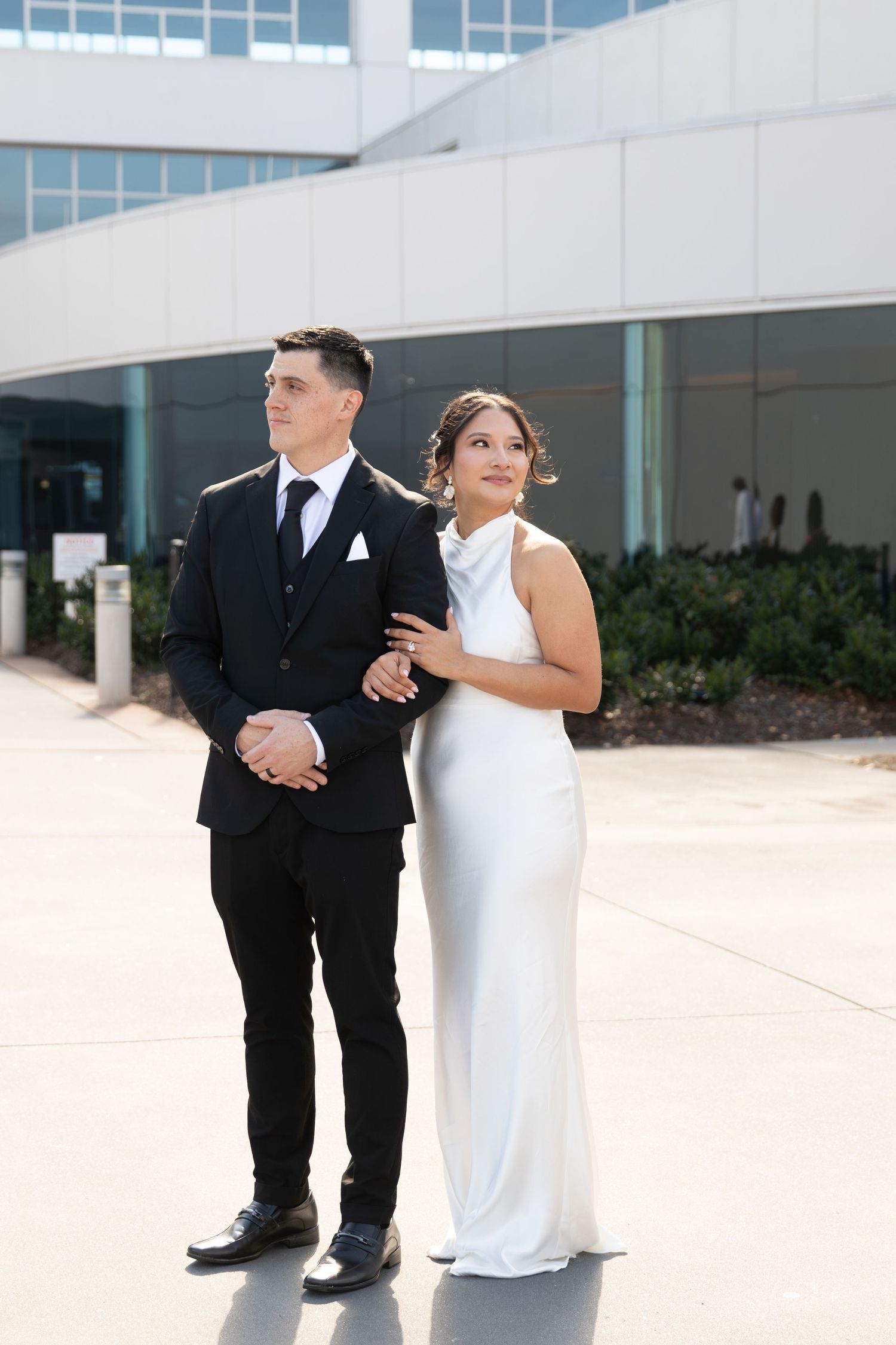 Elegant couple posing outdoors, groom in black suit and bride in white sleeveless gown, in front of modern building.