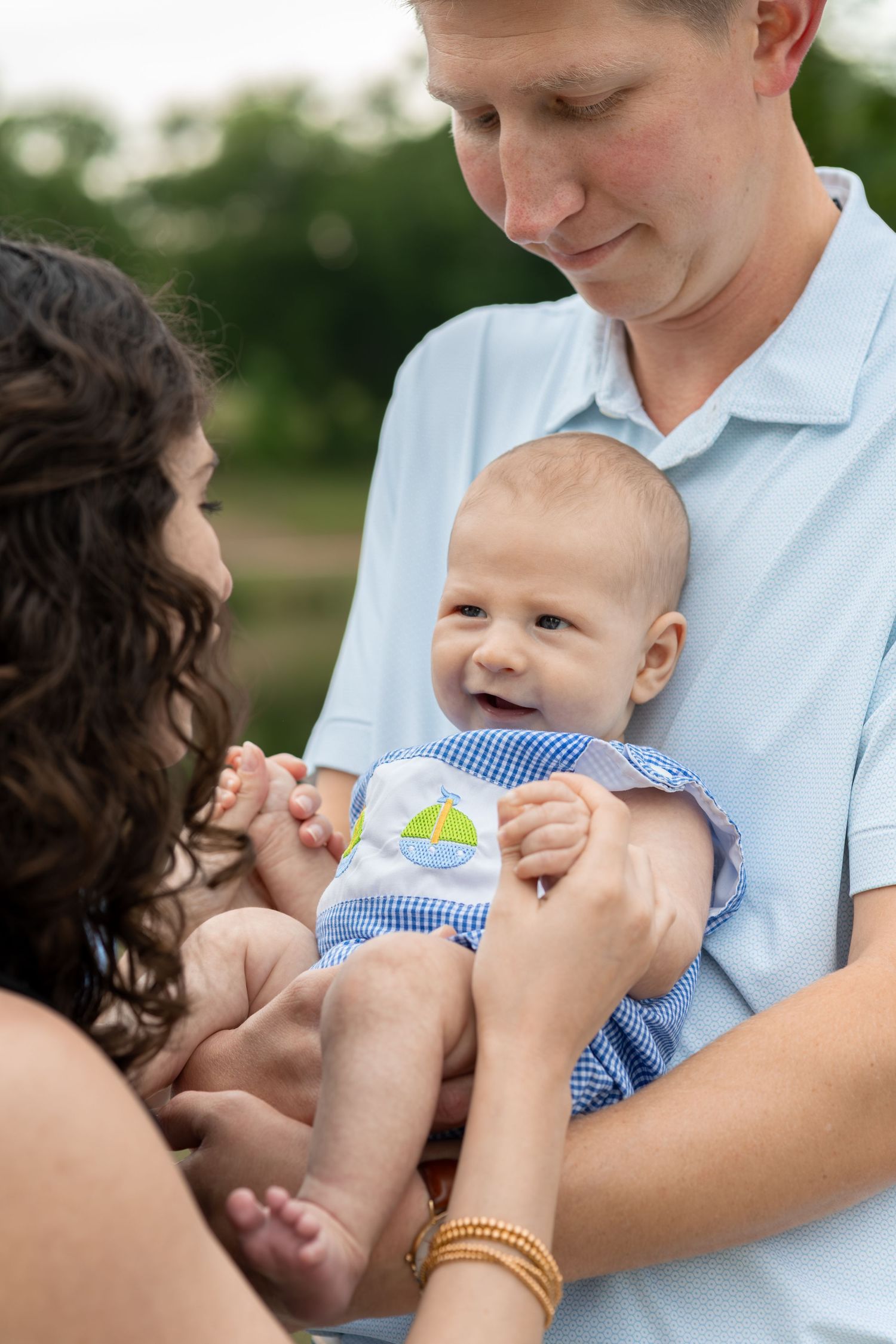 Happy baby in blue gingham outfit smiles while being held by parents outdoors in a green park setting.