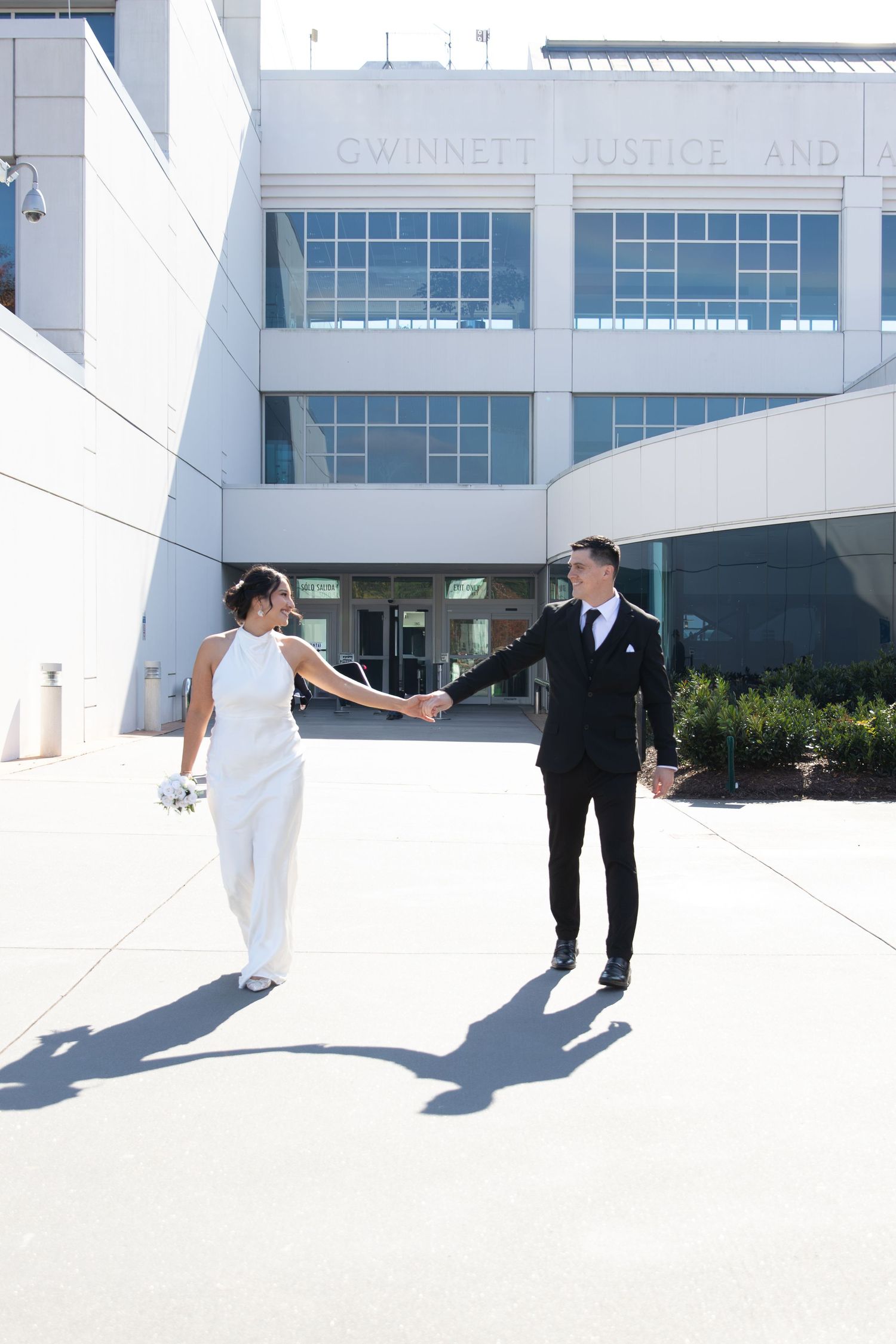 Bride in white jumpsuit and groom in black suit hold hands outside Gwinnett Justice Center on their wedding day.